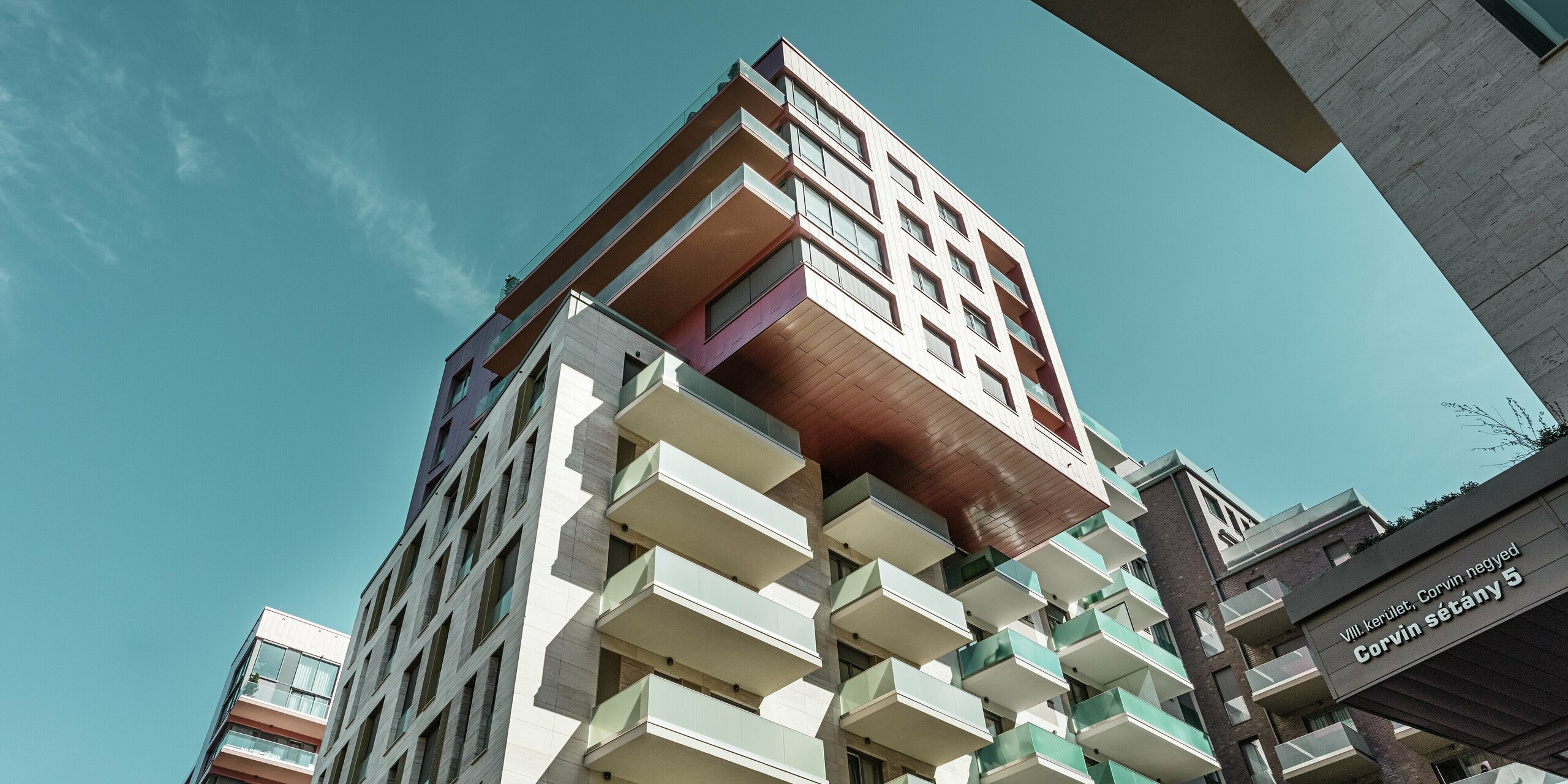 Vue aérienne saisissante du complexe résidentiel Grand Corvin, situé dans le quartier Corvin de Budapest, avec sa partie en porte-à-faux entièrement revêtue de panneaux de façade Siding PREFA d'une couleur saumon métallisé sur-mesure. L'installation verticale de cette façade en aluminium durable confère à ce bâtiment urbain moderne une structure élégante et met en valeur l'architecture de haute qualité dans le cadre d'un vaste programme de rénovation urbaine.
