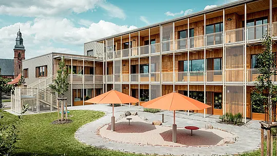 L'arrière de la crèche d'Alzenau dévoile une cour intérieure abritée et son aire de jeux attenante. Deux parasols orange apportent une touche de couleur au-dessus du bac à sable circulaire. À l'arrière-plan, l'aile résidentielle, construite en bois massif durable, se prolonge par un balcon métallique en saillie et un escalier. À gauche, la façade en aluminium est visible, revêtue de losanges de façade 44x44 PREFA couleur bronze – une protection robuste et esthétique contre les intempéries. L'alliance de l'aluminium, du bois et de l'acier symbolise l'intégration intelligente de la fonctionnalité, de la durabilité et de la qualité architecturale. La vue de l'église en arrière-plan ancre le projet dans le paysage urbain d'Alzenau.