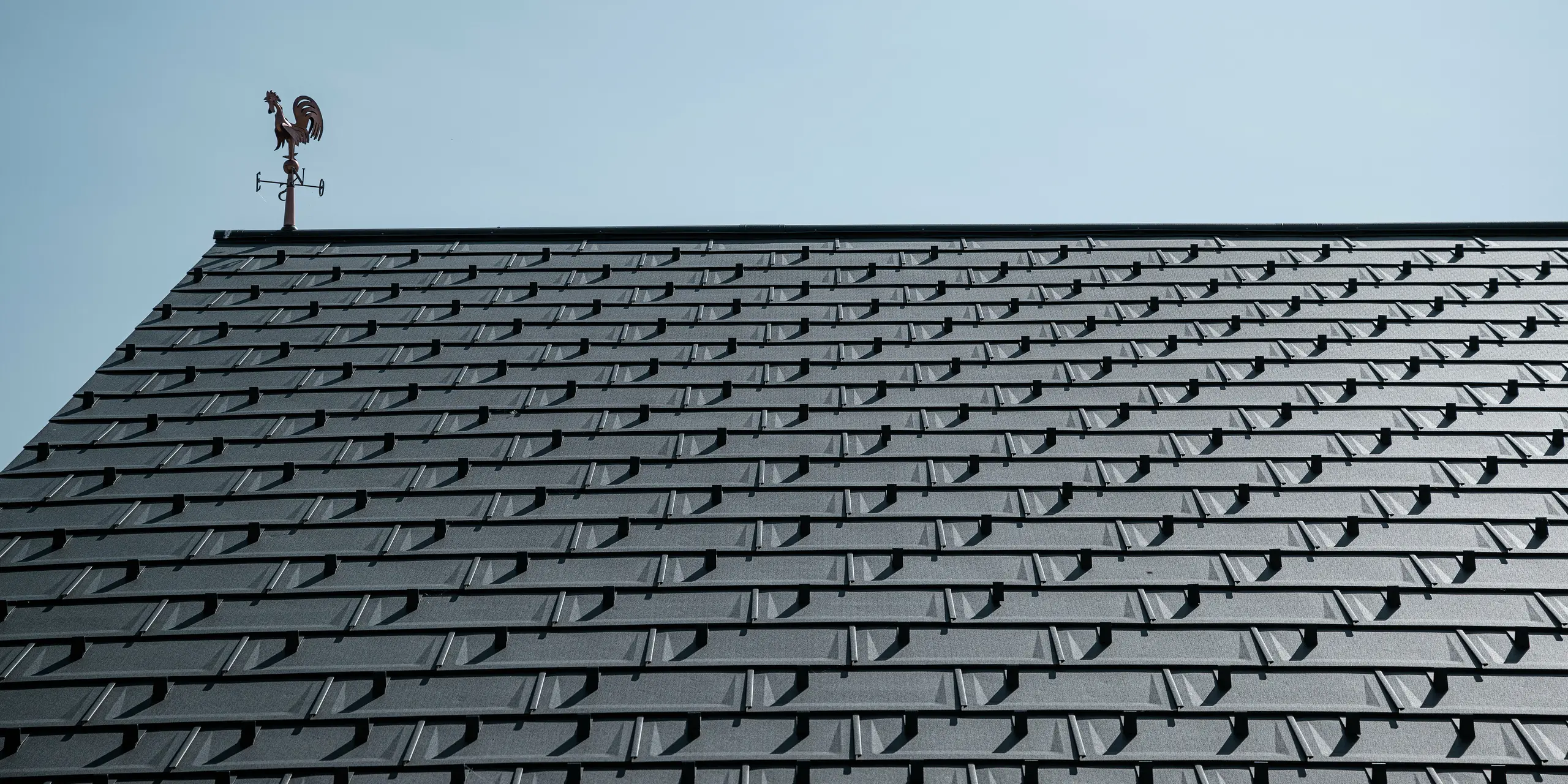 Vue détaillée d'une toiture PREFA avec les R.16 en P.10 gris sombre. L'aspect clair et moderne des panneaux de toiture en aluminium garantit une solution de toit durable, résistante aux intempéries et sans entretien. Les crochets à neige offrent une sécurité supplémentaire dans les régions enneigées. Le toit est couronné d'une girouette classique qui allie tradition et technologie moderne. Grâce aux R.16 robustes PREFA, le toit reste stable et esthétique pendant des décennies.