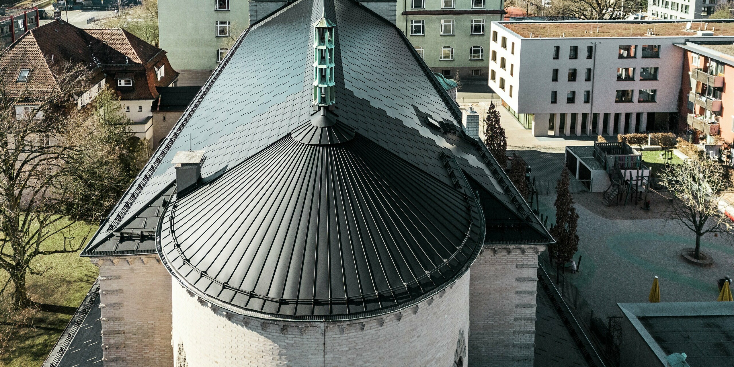 L'image montre le toit arrondi et saisissant à l'arrière de l'église Saint-Charles-Borromée de Nuremberg. La courbe du bâtiment a été recouverte d'une toiture à joint debout en aluminium noir. Le toit à deux versants, qui protège la partie principale du bâtiment, a été recouvert de tuiles solaires PREFA noires. Ces tuiles solaires innovantes constituent un point fort visuel et produisent simultanément de l'électricité à partir d'énergies renouvelables. La vue sur le toit et la flèche centrale illustre l'intégration harmonieuse de la technologie solaire moderne dans l'architecture historique. À l'arrière-plan, on distingue des bâtiments traditionnels et modernes, soulignant la combinaison réussie entre préservation du patrimoine et construction durable.