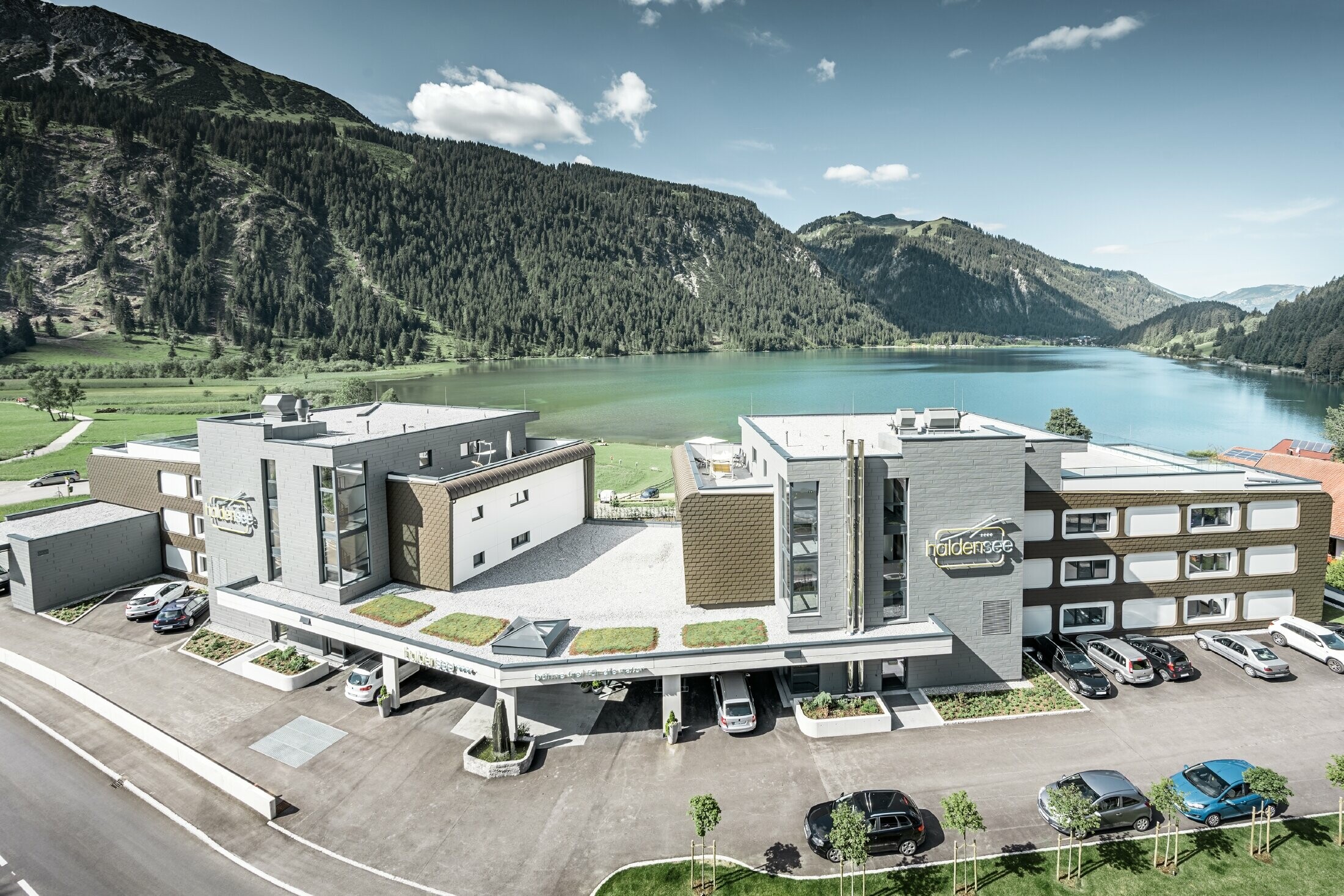 Hotel am Haldensee mit  Aluminium Verbundplatten, Wandschindeln, Prefalz und Siding.X, hinter dem Hotel sind Berge und ein See zu sehen