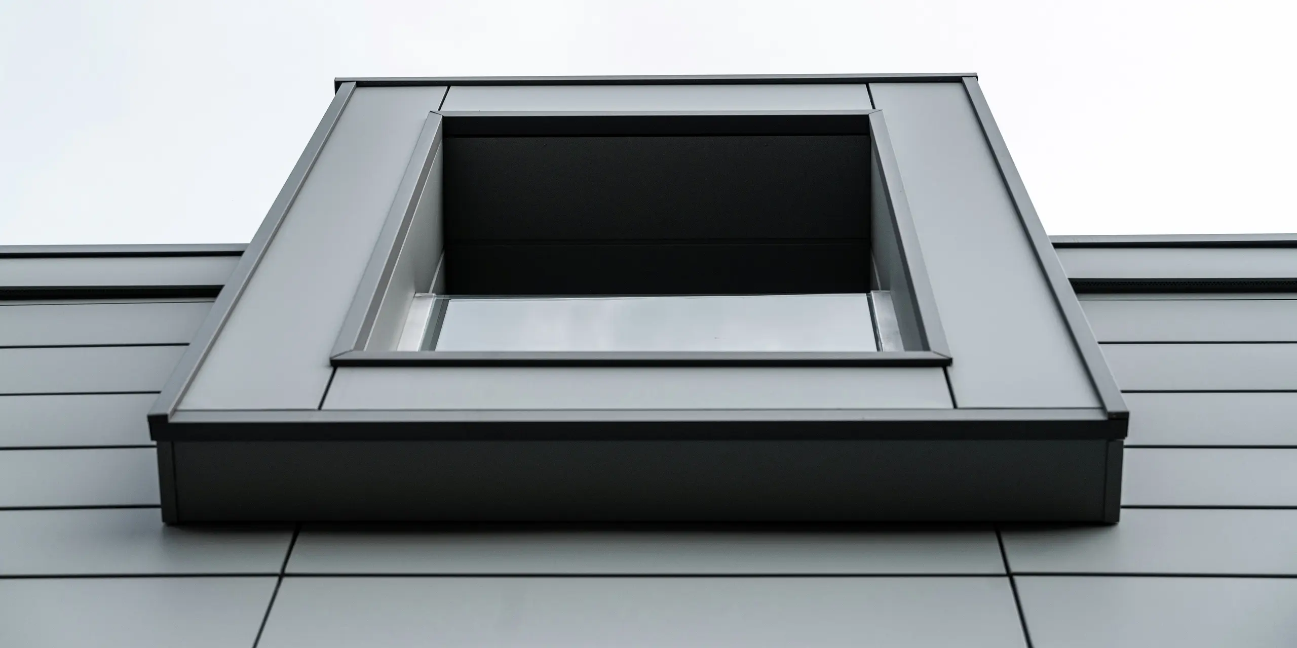 Vue rapprochée d'en bas d'une fenêtre en porte-à-faux d'un complexe résidentiel à Faloppio, en Italie. La forme claire et géométrique de la fenêtre, combinée à la façade en aluminium durable PREFA Siding, souligne l'architecture moderne et le design minimaliste du bâtiment.