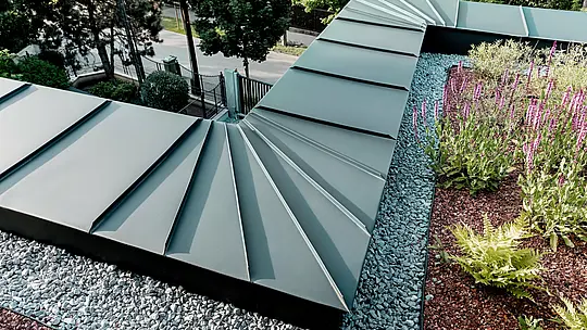 La photographie présente un gros plan détaillé du parapet de la toiture d'une villa multigénérationnelle à Budapest, revêtue d'aluminium PREFALZ couleur P.10 anthracite. La disposition en éventail des panneaux converge précisément au bord du bâtiment, témoignant de la grande formabilité et de la qualité de fabrication des produits en aluminium PREFA. Le métal anthracite offre un contraste élégant avec la toiture végétalisée, agrémentée d'un lit de gravier et de plantes fleuries. Cette scène met en valeur la durabilité, la résistance aux intempéries et la technicité de la solution PREFA.