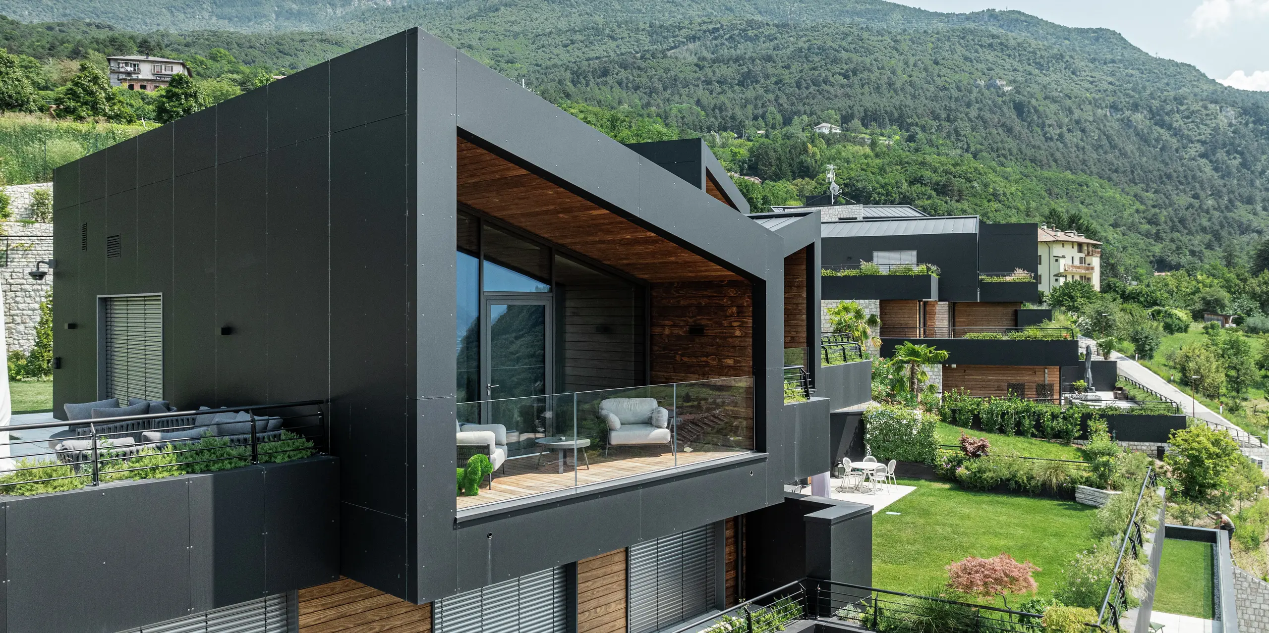 Architecture contemporaine en pleine nature : la généreuse façade vitrée de cet immeuble résidentiel offre une vue imprenable sur le paysage verdoyant et vallonné. La façade en panneaux composites en aluminium PREFA en P.10 Noir crée une enveloppe de bâtiment élégante et résistante aux intempéries qui contraste avec le bois chaleureux. Le design moderne est complété par une terrasse couverte avec des garde-corps en verre, qui relie parfaitement les espaces intérieurs et extérieurs. Grâce aux matériaux PREFA durables et faciles d'entretien, l'apparence reste durablement attrayante - parfaite pour une vie durable avec style.