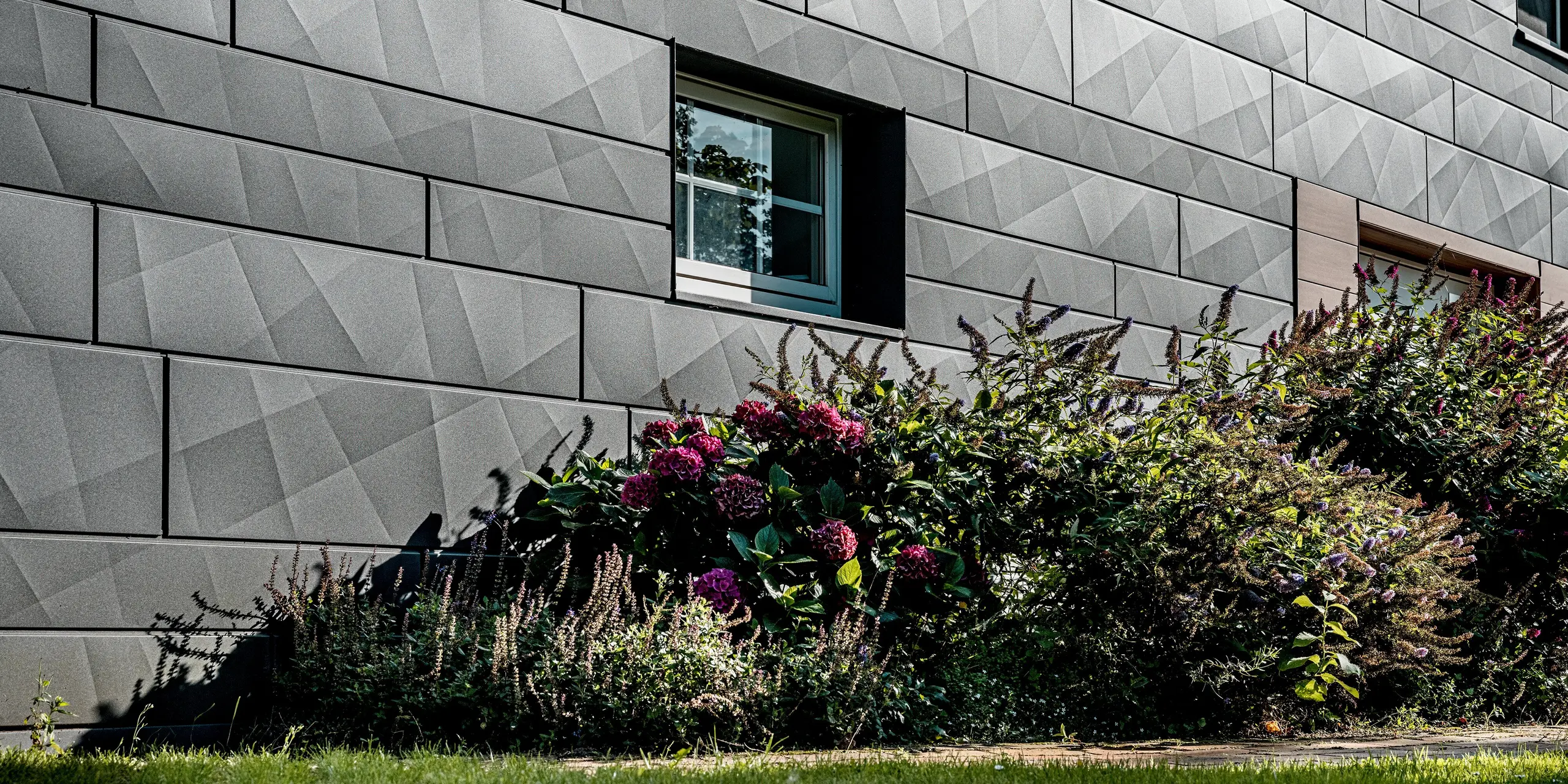 Gros plan sur la façade rénovée d'une maison individuelle revêtue du bardage Siding.X PREFA  en coloris P.10 gris sombre. Les panneaux d'aluminium grand format, aux plis longitudinaux et transversaux marqués, créent une texture géométrique dynamique qui offre un jeu d'ombres et de lumières captivant selon l'angle d'éclairage. Une fenêtre parfaitement ajustée apporte une touche architecturale affirmée et souligne la qualité de la réalisation. Au premier plan, un parterre luxuriant, aux fleurs colorées et à la verdure luxuriante, contraste avec l'élégance sobre de la façade en aluminium. Résistants aux intempéries, inaltérables et faciles d'entretien, les panneaux d'aluminium PREFA allient design expressif et durabilité, et sont idéaux pour les rénovations de façades modernes et écologiques.