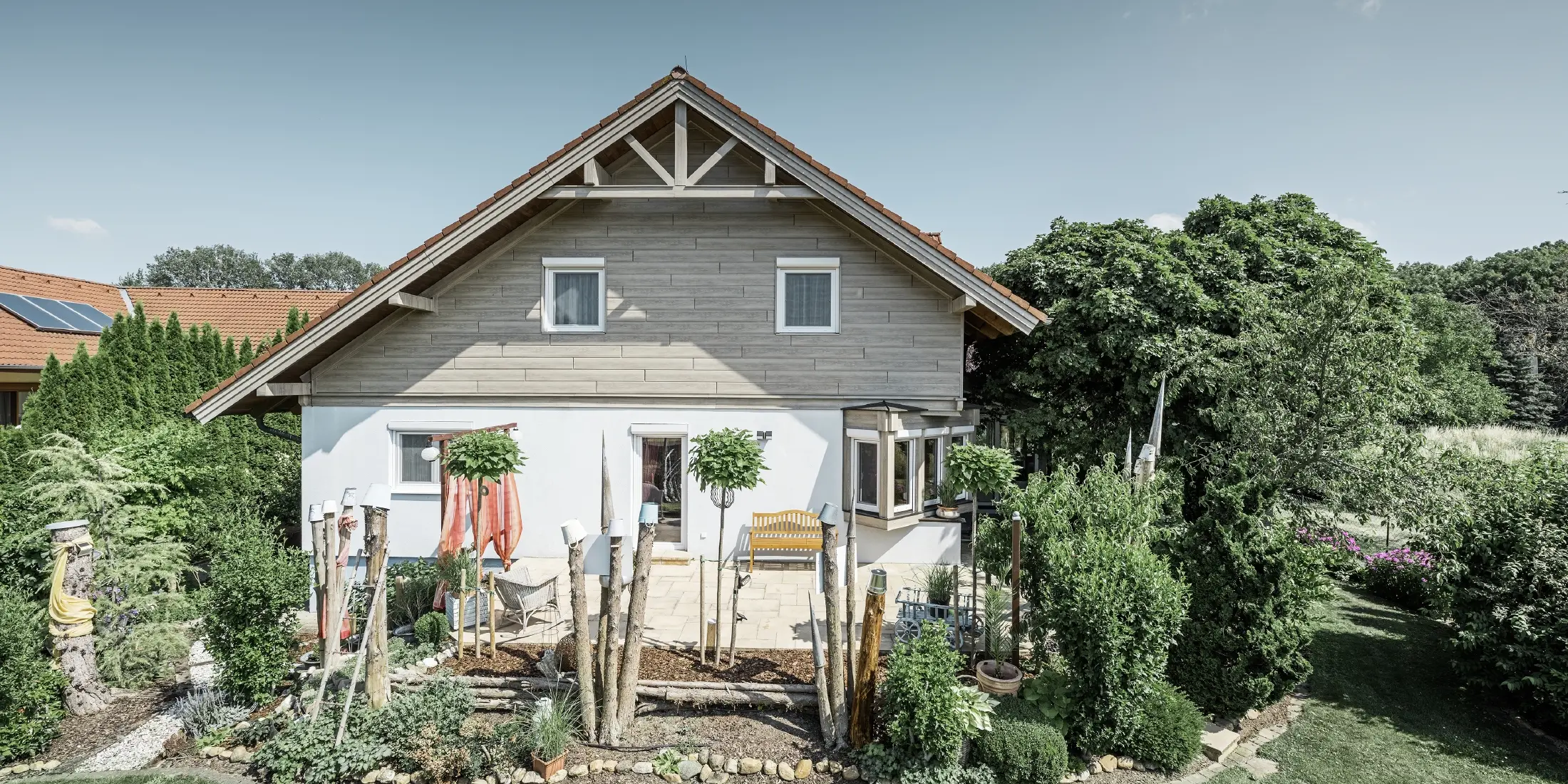 Fassadensaniertes Einfamilienhaus mit erneuerter Giebelverkleidung in moderner Holzoptik in Grau-Beige, kombiniert mit verputzter Fassade, Satteldach und gepflegtem Garten.