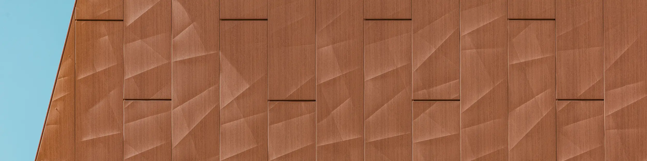 Gros plan sur la façade en aluminium PREFA Siding.X de la station de pompage d'eau potable WML à Lottum. Ses panneaux, pliés avec précision, présentent une patine chaude rappelant la rouille. La structure géométrique de la surface crée un fascinant jeu d'ombres, de lumières et de fines lignes qui se modifie dynamiquement selon l'angle de vue. Les panneaux en aluminium PREFA, de haute qualité, séduisent par leur légèreté, leur grande résistance aux intempéries, leur inaltérable et leur recyclabilité complète, offrant ainsi une solution de façade durable, écologique et nécessitant peu d'entretien.