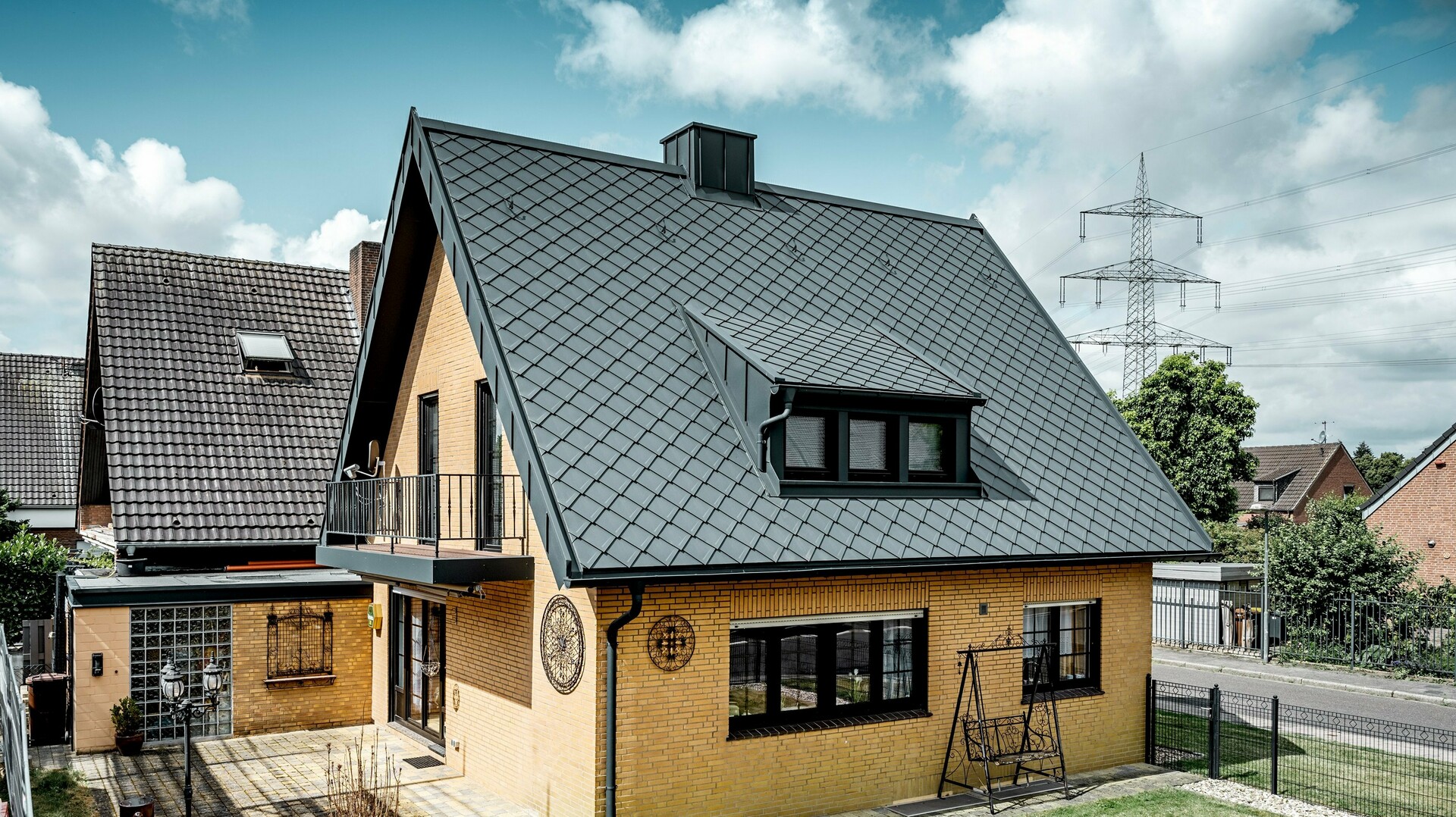 Einfamilienhaus aus gelbem Klinker nach der Dachsanierung mit PREFA Dachraute 29 × 29 in Dunkelgrau; neu eingedecktes, steiles Satteldach mit Rautenmuster, integrierter Gaube und Kamin, Terrasse und Garten im Vordergrund.
