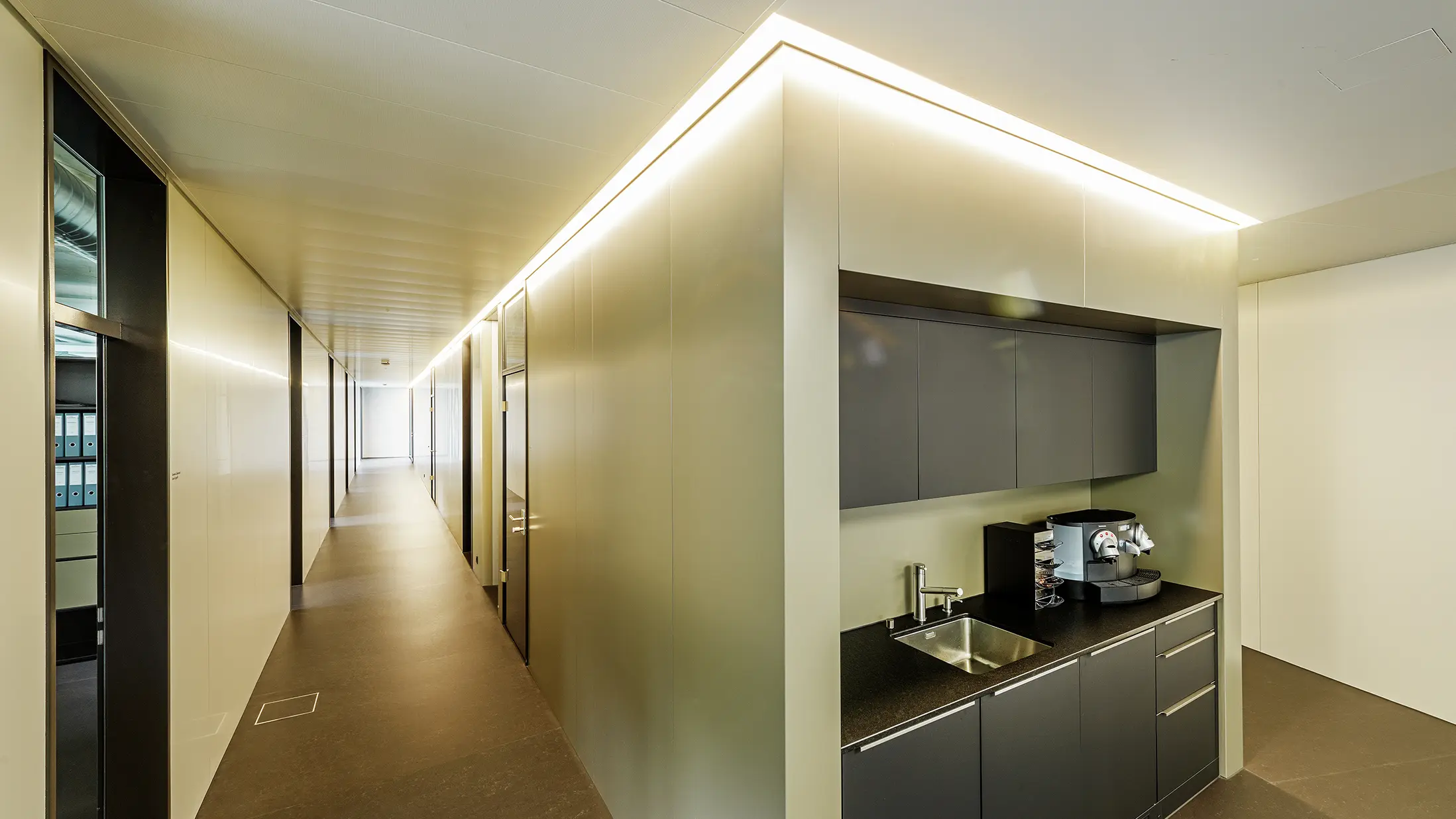 In den modernen Büroräumlichkeiten der Firma Guggisberg Dachtechnik in Wabern sorgt die integrierte Teeküche für Funktionalität und Design. Die Wandverkleidungen nehmen die edle Bronzeoptik der PREFA Aluminiumfassade auf und fügen sich nahtlos in das architektonische Konzept ein. Die Innenraumgestaltung mit indirekter Beleuchtung schafft ein angenehmes Raumgefühl.