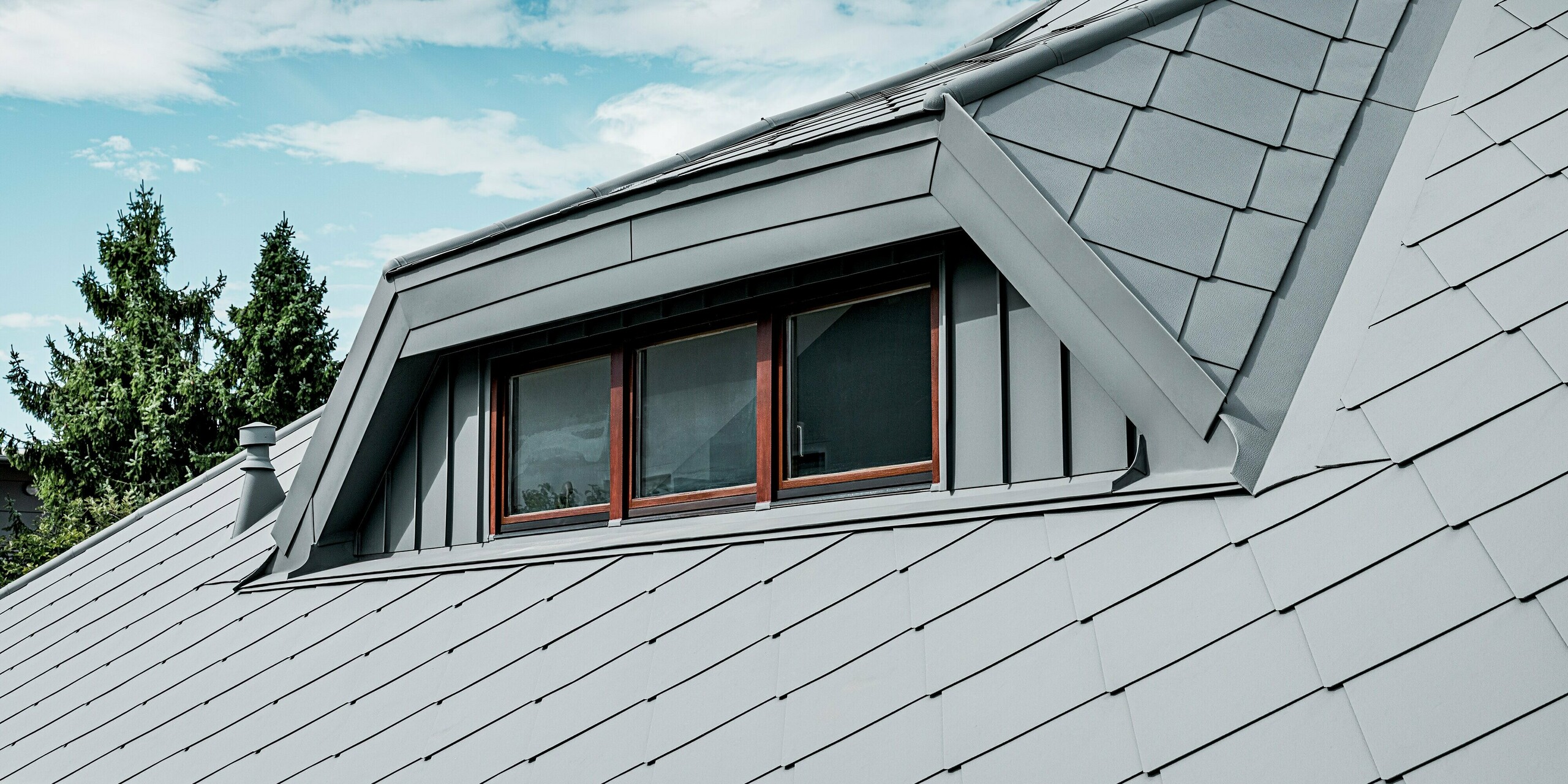 Das Bild zeigt eine Nahaufnahme der markanten Trapezgaube eines Einfamilienhauses, deren Oberfläche vollständig mit PREFA Aluminium in P.10 Hellgrau gestaltet ist. Die präzise integrierte Dachraute 44 × 44 und die klar definierten Kanten der Gaubenkonstruktion erzeugen eine moderne, hochwertige Optik. Die feine Stucco-Struktur verleiht dem Aluminiumdach zusätzliche Tiefe und Robustheit. Unter dem hellen Himmel wirkt das langlebige, witterungsbeständige PREFA Dachsystem besonders elegant und stabil.