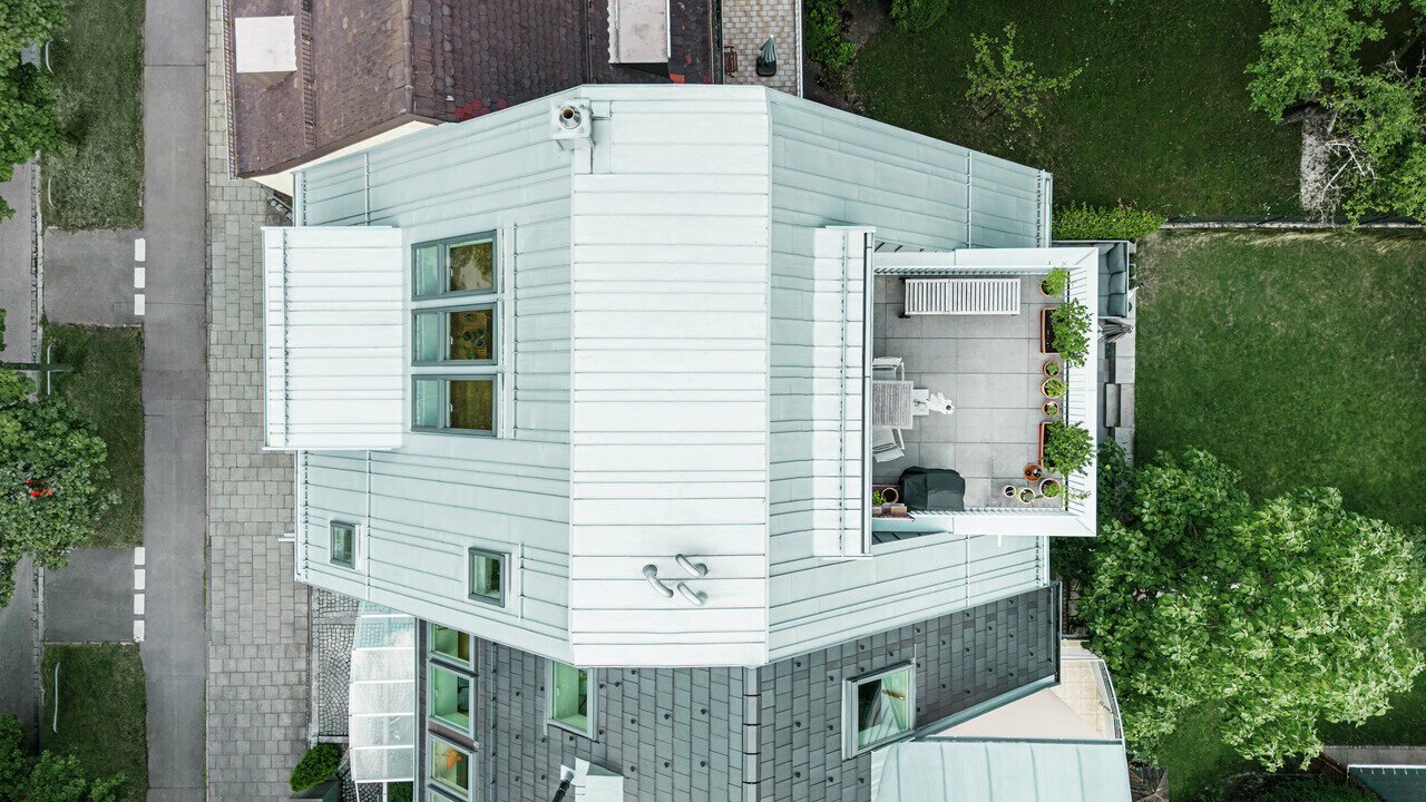 Luftaufnahme eines Eckreihenhauses mit moderner Sanierung. Das Haus verfügt über ein Dach und eine Obergeschossfassade aus silbermetallischem Prefalz Aluminium, die horizontale Linienführung betont. Eine Dachterrasse mit Outdoor-Möbeln und Pflanzen schafft eine urbane grüne Oase. Die Untergeschossfassade ist in einem dunkleren Ton gehalten, was einen ansprechenden Kontrast bildet. Umgeben ist das Haus von einer gepflegten Grünfläche und gepflasterten Wegen, die die klare Struktur des Gebäudes unterstreichen.