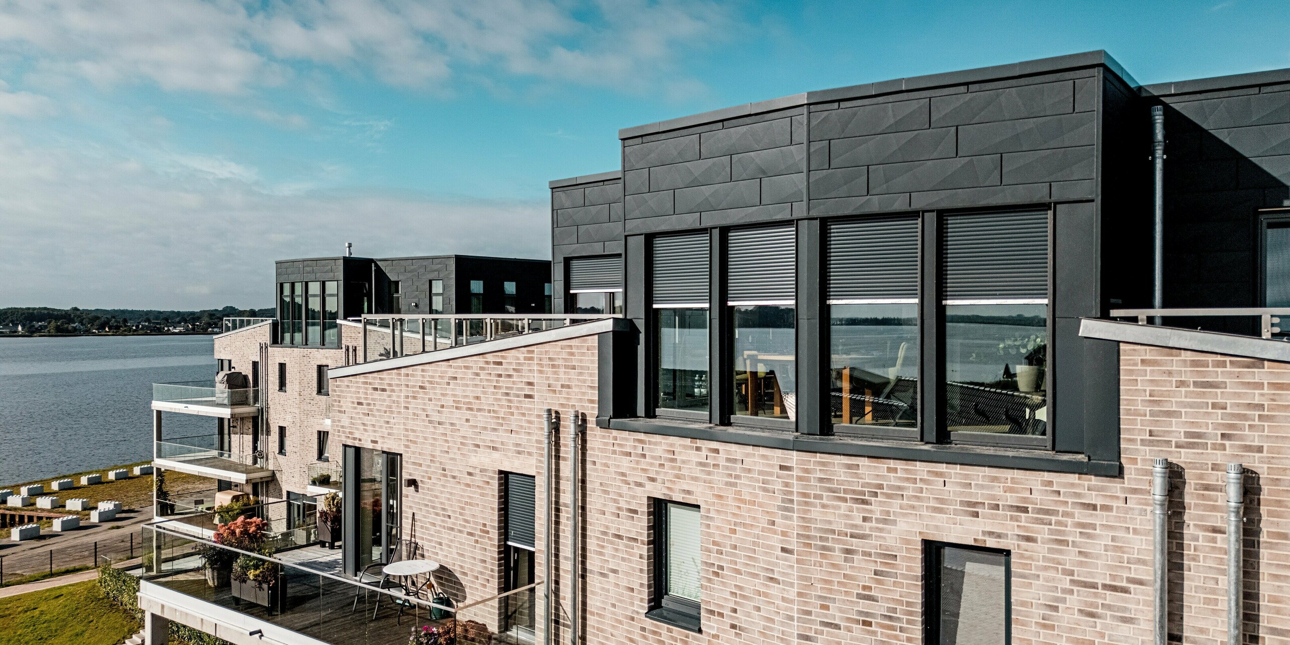 Das Luftbild zeigt die Wohnanlage „Hafen zur Freiheit“ direkt am Wasser, wobei das markante Dachgeschoss mit PREFA Siding.X in P.10 Anthrazit besonders ins Auge fällt. Die dunkel strukturierten Aluminium­paneele setzen einen starken Kontrast zur hellen Klinkerfassade und verleihen dem Gebäude eine moderne, robuste Anmutung. Großzügige Fenster und offene Terrassen verstärken die offene, lichtdurchflutete Architektur. Dank der widerstandsfähigen PREFA Materialien bleibt die Fassade selbst in der exponierten Küstenlage dauerhaft geschützt und optisch hochwertig.