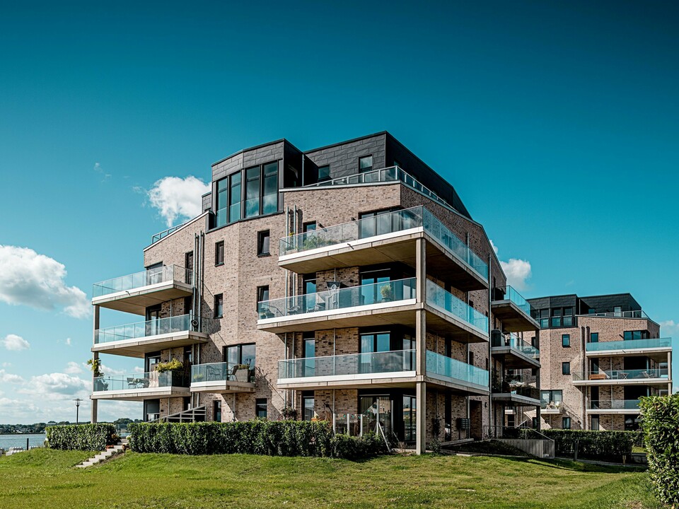 Die Wohnhausanlage „Hafen zur Freiheit“ in Schleswig präsentiert moderne Architektur direkt am Wasser: helle Klinkerfassaden treffen auf dynamisch strukturierte PREFA Siding.X Elemente in P.10 Anthrazit. Besonders auffällig sind die oberen Geschosse mit großzügigen Glasbalustraden und klaren Linien, die im Sonnenlicht elegant kontrastieren. Die widerstandsfähigen Aluminiumfassaden sorgen für langlebigen Schutz vor Küstenwetter und verleihen dem Ensemble einen markanten, hochwertigen Charakter. Die großzügigen Balkone öffnen den Blick auf die maritime Umgebung und unterstreichen das moderne Wohngefühl.