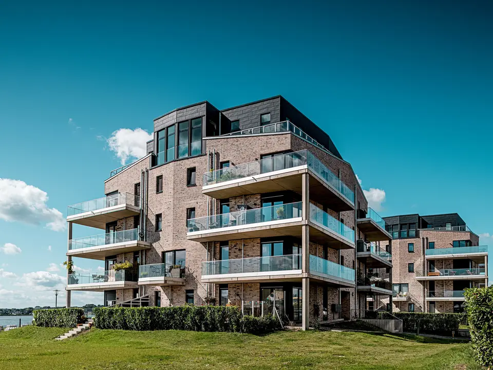 Le complexe résidentiel « Hafen zur Freiheit » (Port vers la liberté) à Schleswig présente une architecture moderne en bord de mer : des façades en briques claires côtoient des éléments Siding.X PREFA P.10 anthracite à la structure dynamique. Les étages supérieurs, particulièrement remarquables, se distinguent par leurs généreuses balustrades en verre et leurs lignes épurées qui créent un élégant contraste sous la lumière du soleil. Les façades en aluminium résistant assurent une protection durable contre les intempéries côtières et confèrent au complexe un caractère distinctif et haut de gamme. Les balcons spacieux offrent une vue imprenable sur le paysage maritime et contribuent à un confort de vie optimal.