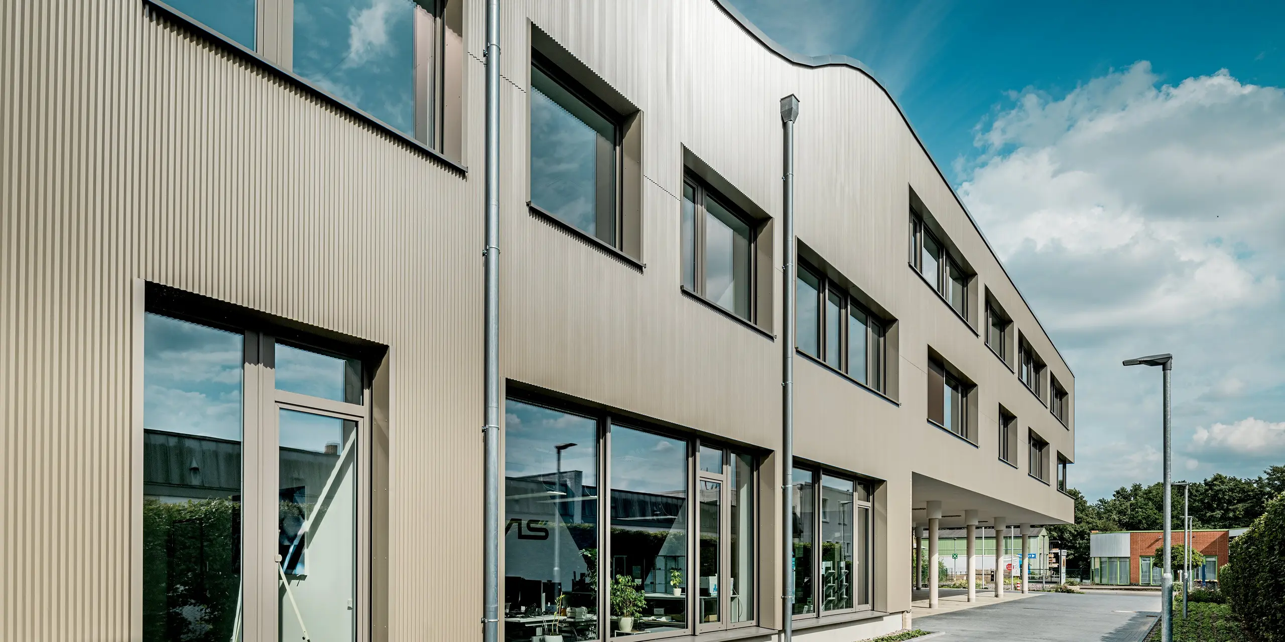 L'image présente le bâtiment moderne Festo à Langenhagen, dont la façade remarquable est composée de profilés extrudés triangle PREFA. Ces dentelures confèrent à la façade une texture distinctive et créent un effet visuel unique. La structure ondulée du bâtiment contraste de manière saisissante avec le design géométrique épuré et s'intègre harmonieusement à son environnement. La large surface vitrée et les élégants panneaux composites en aluminium complètent parfaitement le design de la façade, offrant une esthétique moderne et intemporelle. Grâce à la durabilité et à la facilité d'entretien des matériaux PREFA, la façade est parfaitement protégée des intempéries et restera en parfait état pendant de nombreuses années.