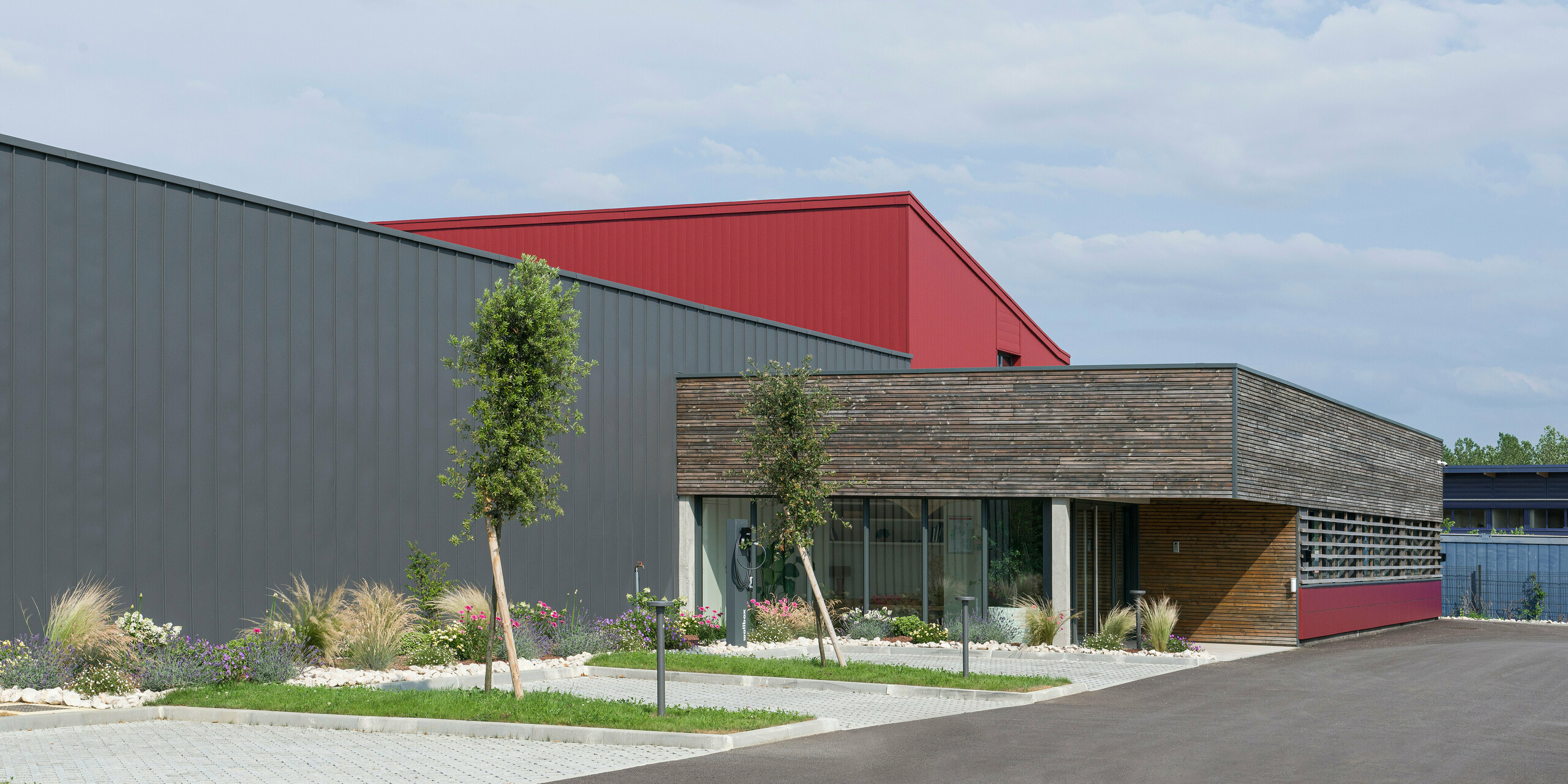  Das Bild zeigt ein modernes Gebäude mit einer auffälligen Kombination aus industriellen und natürlichen Designelementen. Die Fassade kombiniert dunkelgraue und oxydrote Aluminiumelemente, die Dynamik und Struktur verleihen. Die oxydroten PREFA Sidings setzen dabei einen farblichen Akzent. Der niedrigere Gebäudeteil mit einer holzverkleideten Wand und großen Fensterflächen bringt eine warme, einladende Atmosphäre ins Spiel, was auf einen büronutzigen Bereich hindeutet. Der gepflegte Gartenbereich vor dem Gebäude vermittelt ein umweltbewusstes und nachhaltiges Image. Die Kombination dieser Elemente spiegelt das Bestreben wider, traditionelle Bauweisen mit modernen, nachhaltigen Materialien und Designs zu verbinden.