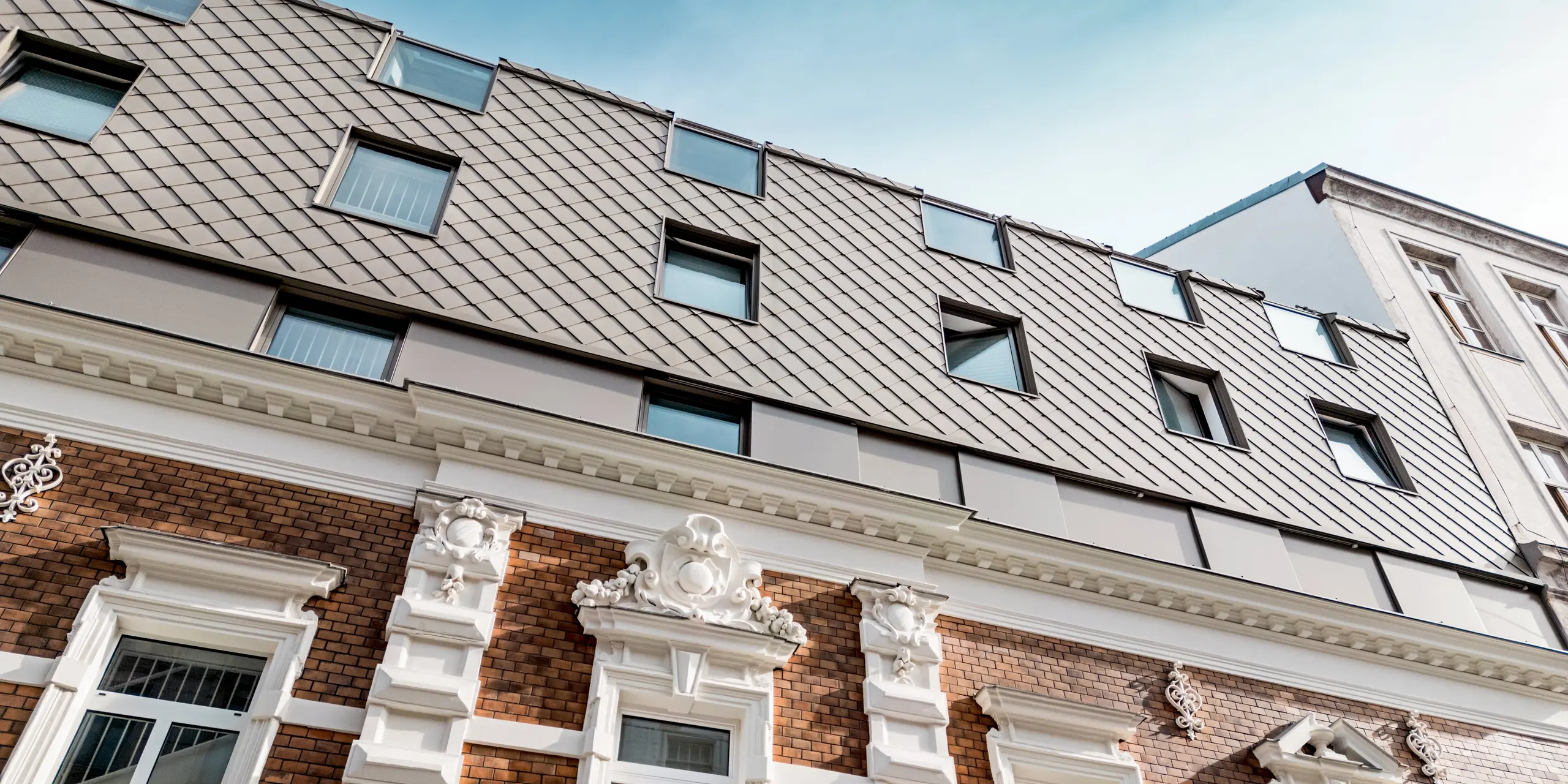 L'image illustre la fusion réussie d'architecture historique et moderne dans un immeuble résidentiel viennois. Les éléments classiques de la façade, avec ses magnifiques stucs et ses briques rouges, sont élégamment mis en valeur par les losanges 29x29 PREFA dans la teinte bronze, qui habillent la toiture. Les fenêtres, judicieusement positionnées à l'étage, s'intègrent harmonieusement à l'ensemble, tandis que la charpente du toit, avec ses losanges, y apporte une touche contemporaine. Cette alliance de tradition et d'innovation confère au bâtiment une allure unique et d'une grande beauté.
