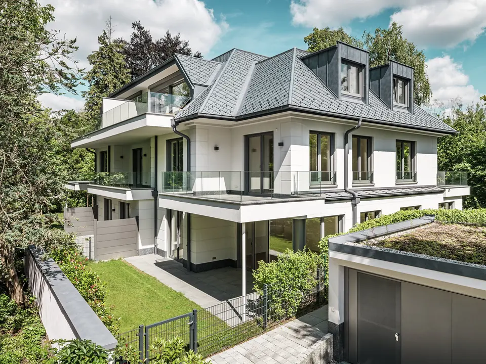 Vue d'un immeuble d'appartements moderne au design élégant près de Munich. La toiture est recouverte de losanges PREFA 29 x 29 cm, coloris Gris Pierre P.10. La charpente remarquable est mise en valeur par d'élégantes lucarnes plates en PREFALZ, à la fois esthétiques et fonctionnelles. Le jardin soigné et la vaste terrasse soulignent la qualité architecturale et créent une harmonie entre intérieur et extérieur. Résistants et durables, les produits PREFA s'intègrent parfaitement au paysage verdoyant et offrent une protection et une beauté pérennes.