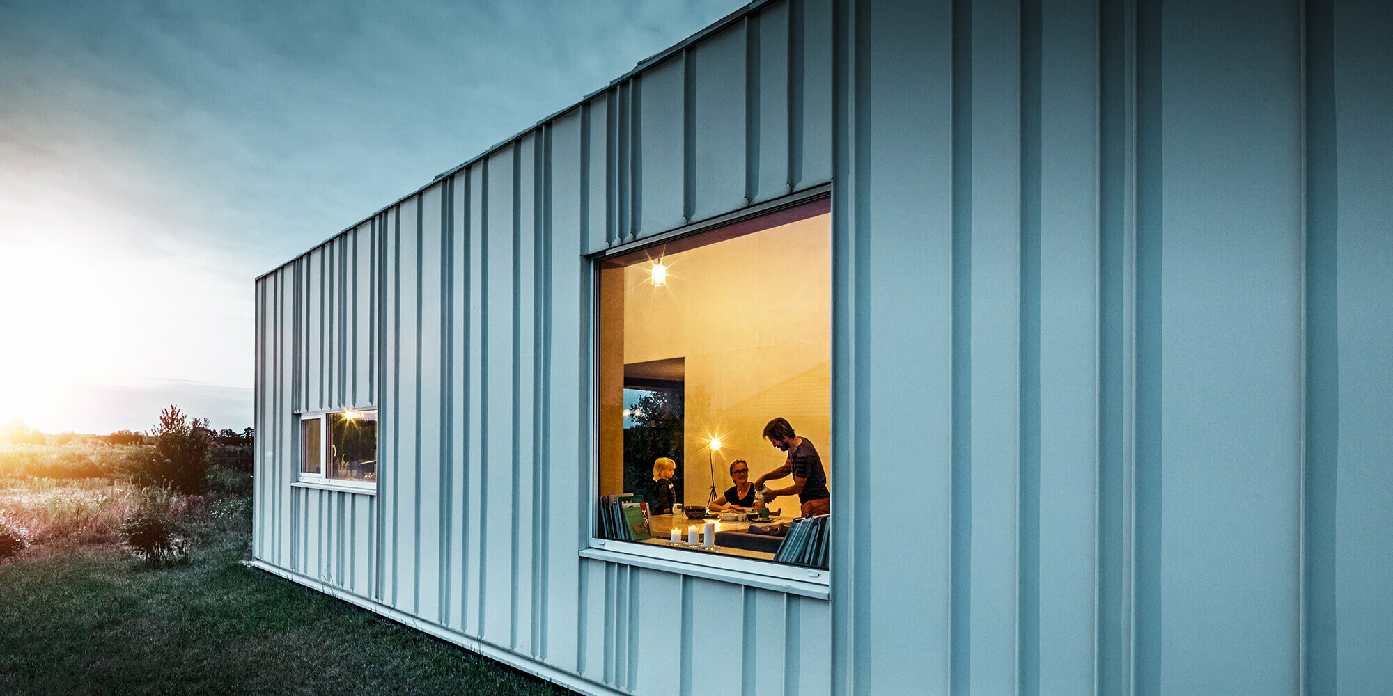 Winkelstehfalz Fassade mit PREFALZ in unterschiedlichen Scharenbreiten in P.10 Prefaweiß bei Dämmerung fotografiert. Durch das Esszimmer erkennt man eine Familie beim Abendessen. 
