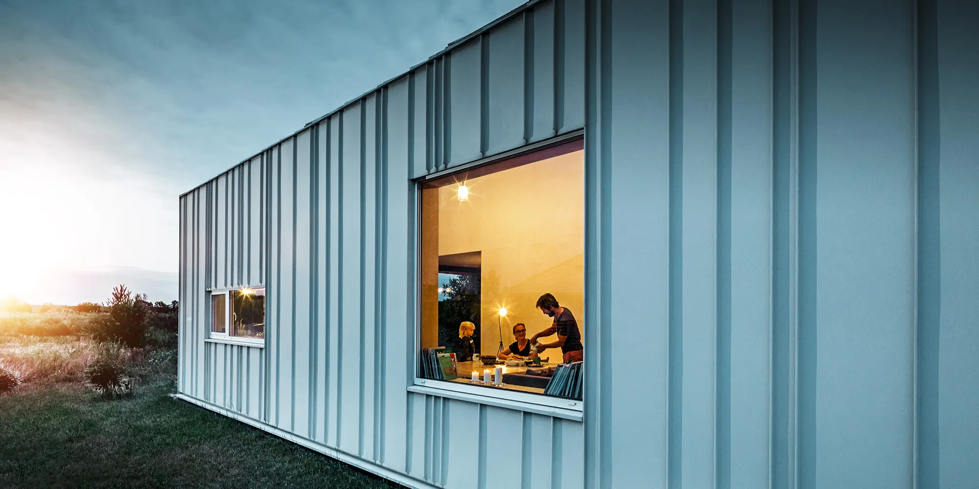 Façade avec joint debout et différentes largeurs de lames en PREFALZ de P.10 blanc PREFA photographiée au crépuscule. À travers la fenêtre de la salle à manger, on peut voir une famille en train de dîner.
