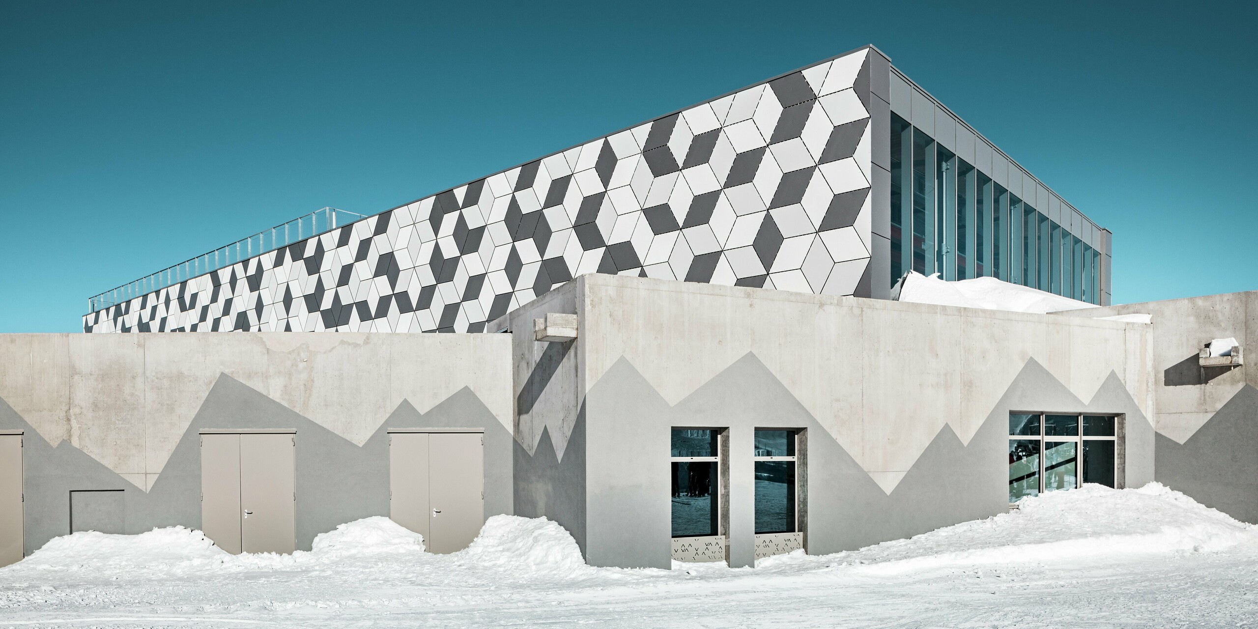 La station d'altitude moderne du téléphérique 3S des Deux Alpes arbore une façade futuriste en panneaux composites en aluminium PREFABOND au motif losange saisissant. Les couleurs P.10 Blanc Prefa, P.10 Blanc Pur et Argent Fumé créent un motif 3D fascinant qui s'intègre parfaitement au paysage enneigé de haute montagne. Résistante aux intempéries et durable, cette façade est un véritable atout esthétique. La partie inférieure du bâtiment est en béton apparent. Les montagnes y sont peintes en gris foncé.