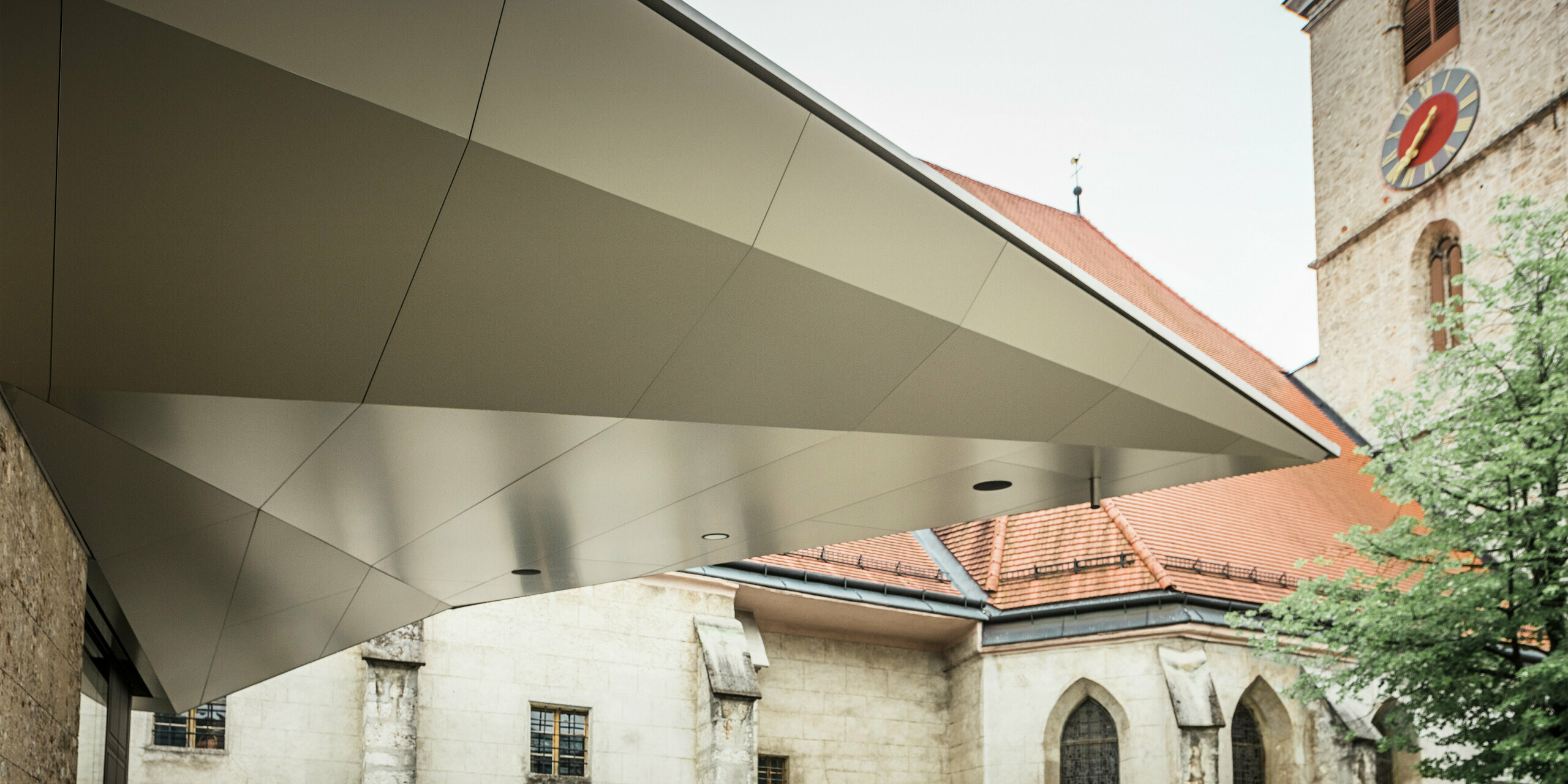 Dynamisch gespannte Dachfläche mit scharfkantiger Linienführung: PREFA Aluminium Verbundplatten ermöglichen diese präzise, fugenarme Gestaltung, die dem Eingangsbereich Leichtigkeit und Modernität verleiht - bei gleichzeitig hoher Witterungsbeständigkeit. Im Hintergrund ist die historische Kirche aus dem 14. Jahrhundert zu erkennen.