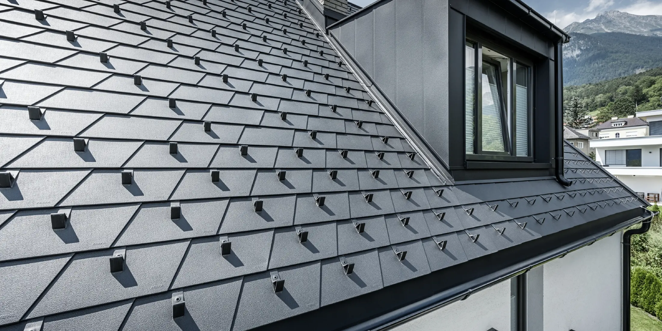Vue détaillée d'une toiture avec lucarne et fenêtre, la surface du toit est recouverte de bardeaux PREFA noirs avec arrêts de neige.