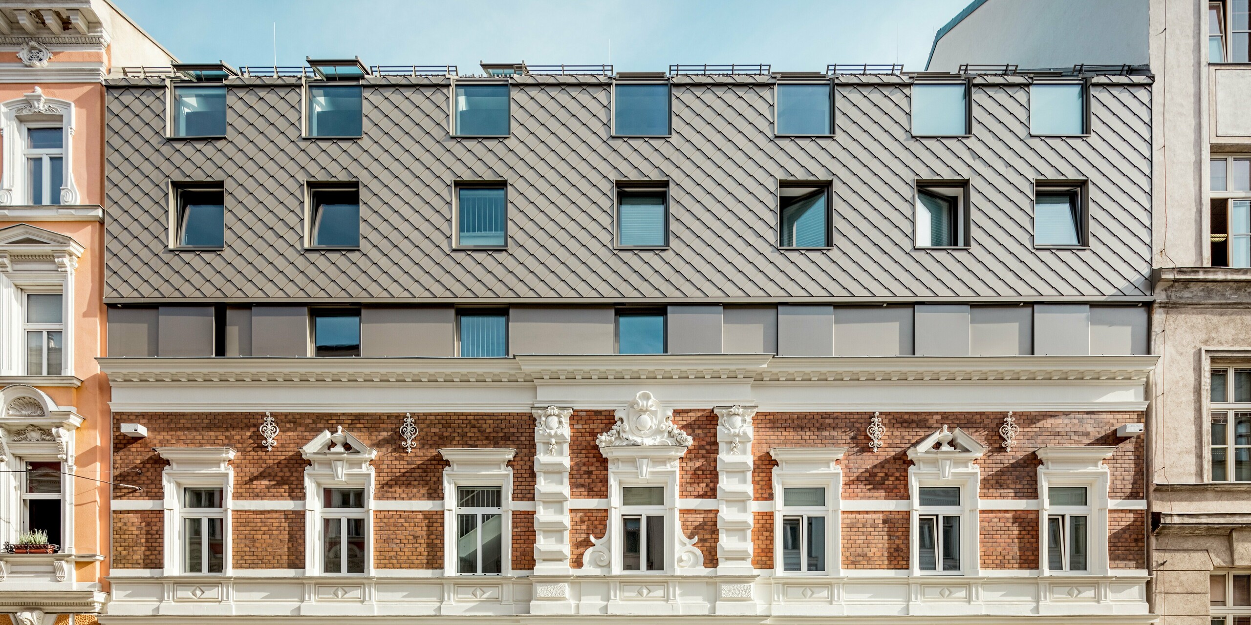 Beeindruckende Fassadenansicht eines modern erweiterten Wohngebäudes in Wien. Das obere Stockwerk wurde mit PREFA Dach- und Wandrauten in einer warmen Bronzeoptik verkleidet, die sich harmonisch in die Architektur der angrenzenden historischen Gebäude einfügt. Besonders auffällig sind die klaren Linien der modernen Fenster und die Struktur der Rauten, die an traditionelle Dachziegel erinnern und dennoch einen zeitgemäßen Akzent setzen. Diese gelungene Kombination von Alt und Neu verleiht dem Gebäude eine einzigartige visuelle Identität.