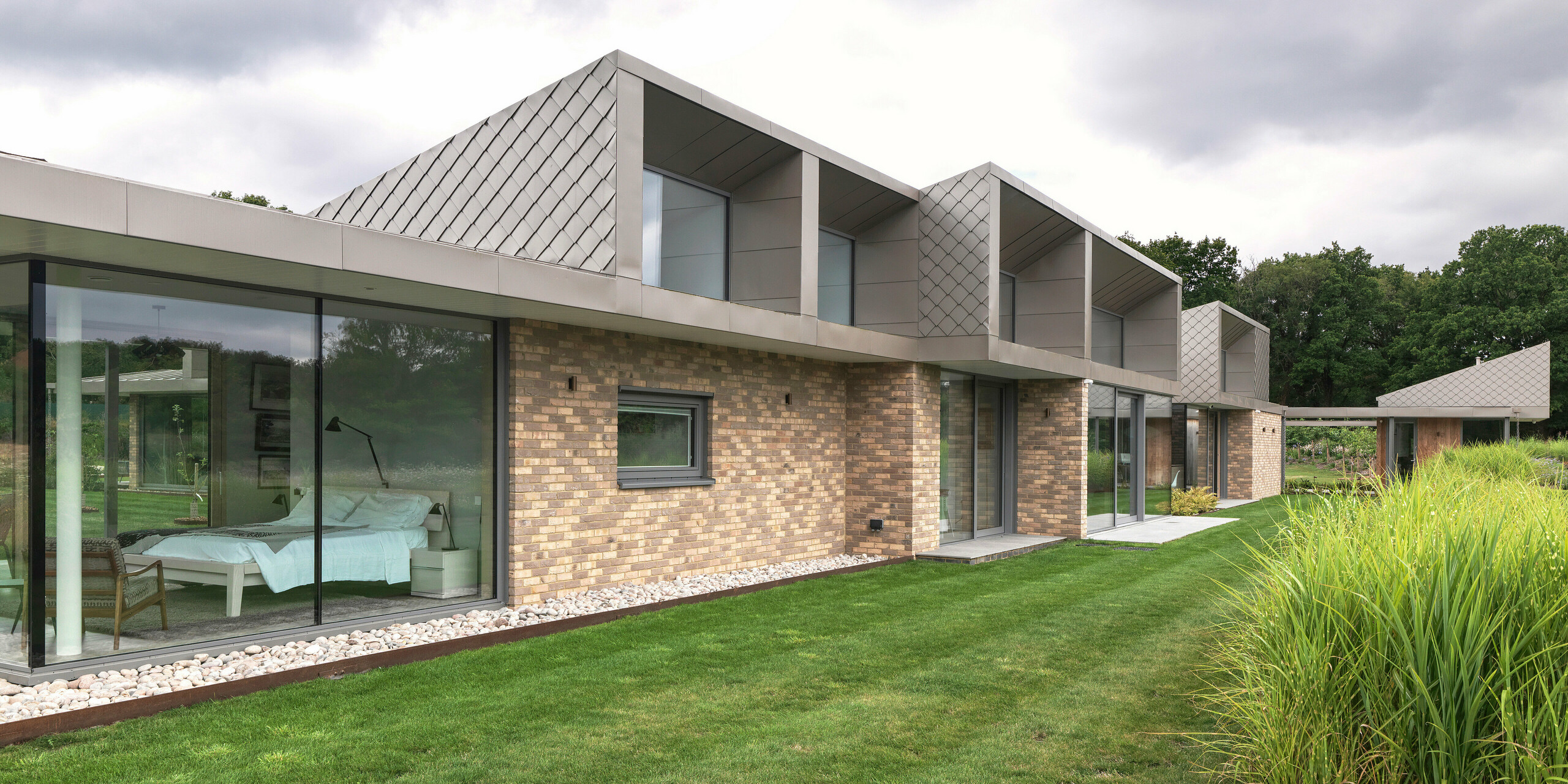 Vue latérale d'un immeuble résidentiel moderne dans le West Sussex, avec ses structures remarquables à l'étage, revêtu d'un bardage en losanges PREFA 20 × 20 en couleur P.10 bronze. La toiture en aluminium PREFALZ, légèrement inclinée, offre une finition élégante et résistante aux intempéries. Au rez-de-chaussée, de larges baies vitrées relient harmonieusement les espaces intérieurs et extérieurs, conférant au bâtiment une allure haut de gamme et intemporelle.