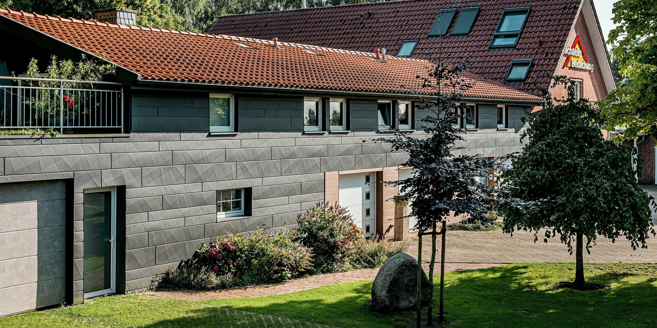 Außenansicht eines sanierten Einfamilienhauses mit moderner PREFA Aluminiumfassade, eingebettet in einen gepflegten Vorgarten mit Bäumen, Sträuchern und blühenden Beeten. Die Fassade kombiniert PREFA Siding.X in P.10 Dunkelgrau im oberen Bereich mit PREFA Siding in Walnussbraun rund um Eingänge und Garagentore. Besonders auffällig sind die markanten Kantungen der dunklen Aluminiumpaneele, die je nach Sonneneinstrahlung ein lebendiges Licht- und Schattenspiel erzeugen. Die warme Holzoptik bildet einen harmonischen Kontrast zur kühlen Eleganz des Aluminiums. Langlebig, witterungsbeständig und pflegeleicht – die PREFA Fassadenlösung verbindet nachhaltige Funktionalität mit hochwertiger Architektur.
