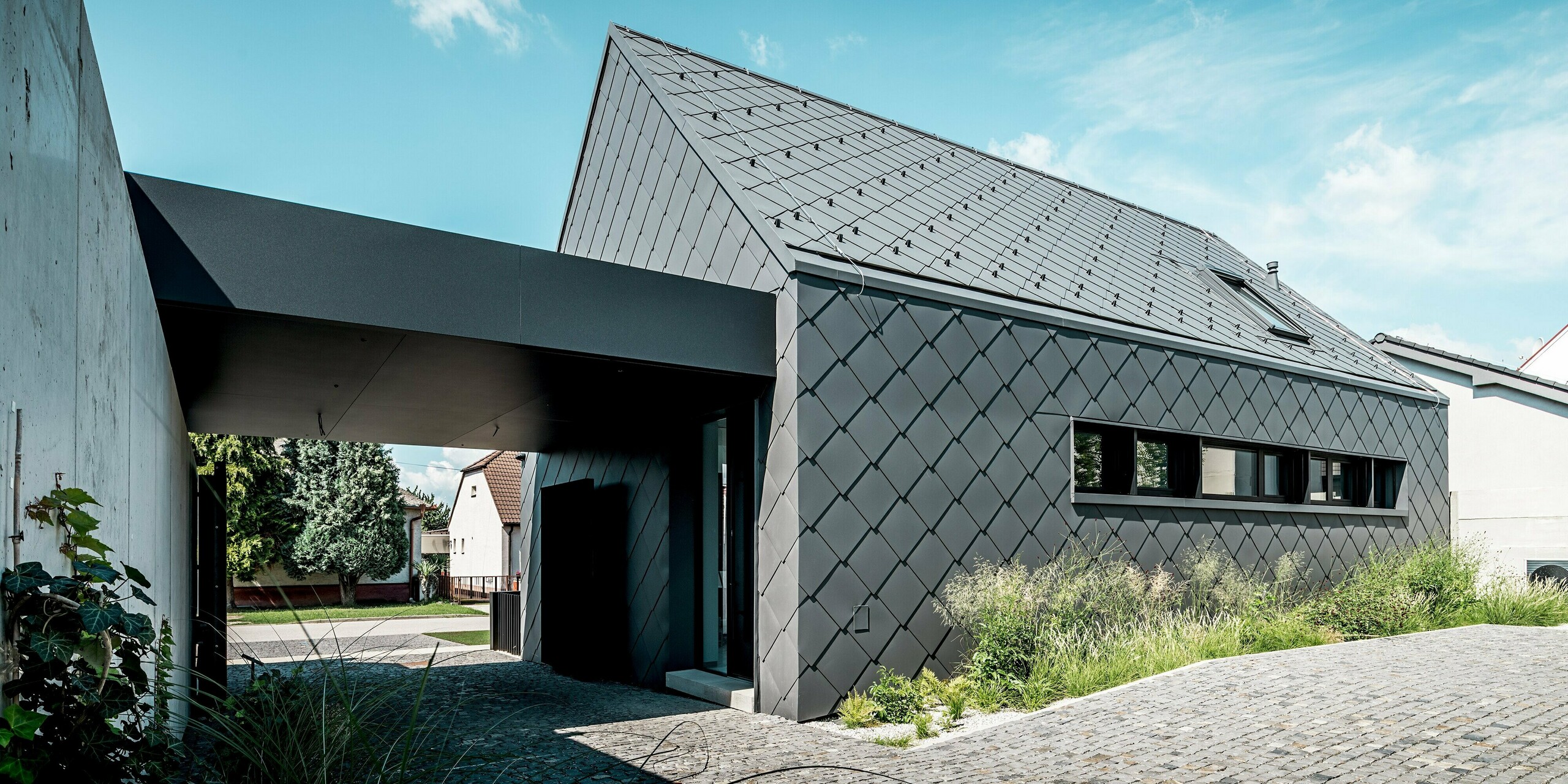 Das Bild zeigt die moderne Architektur eines Einfamilienhauses in Trnava‑Modranka, komplett gestaltet mit PREFA Dach- und Wandrauten 44 × 44 in P.10 Dunkelgrau. Die durchgehende Rautenstruktur betont die klaren Geometrien des Baukörpers und verleiht dem Gebäude eine elegante, monolithische Optik. Die langlebigen Aluminiumprodukte schützen dauerhaft vor Witterung und sorgen für eine hochwertige, wartungsarme Gebäudehülle. Der lichtdurchflutete Vorbereich und die umgebende Begrünung setzen natürliche Kontraste zur markanten PREFA Fassade.