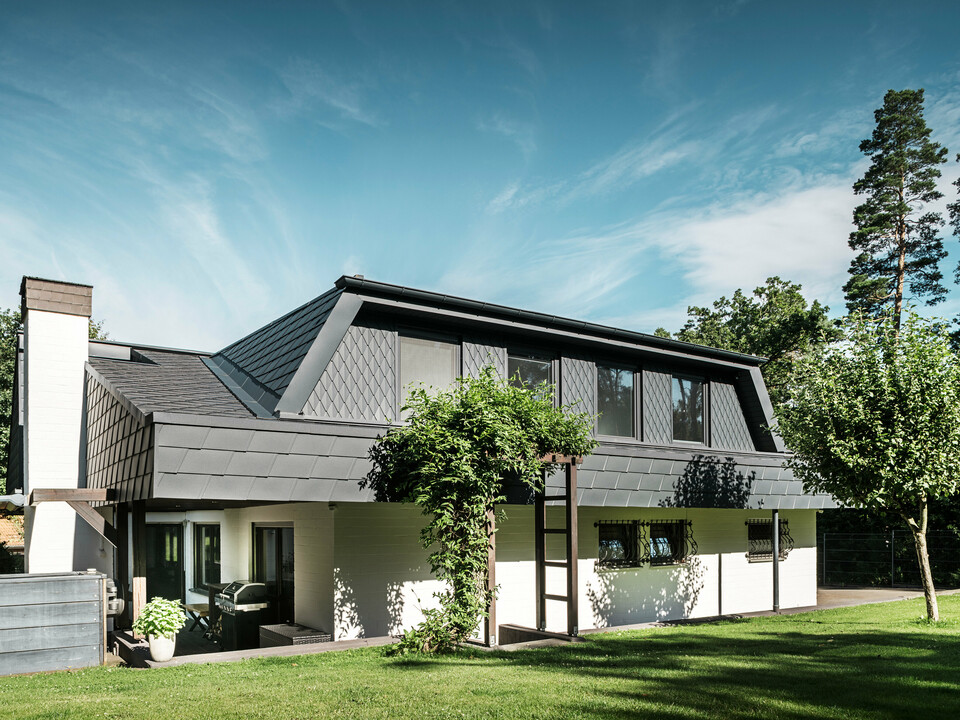 Vue latérale d'une maison individuelle à Mechernich avec des bardeaux DS.19 PREFA et de petits losanges de toiture en anthracite P.10. Les lucarnes trapézoïdales sont recouvertes de losanges de toit en tôle robuste, tandis que le toit est recouvert de bardeaux. Une pergola verte et des arbres dans le jardin complètent l'architecture moderne et résistante aux intempéries.
