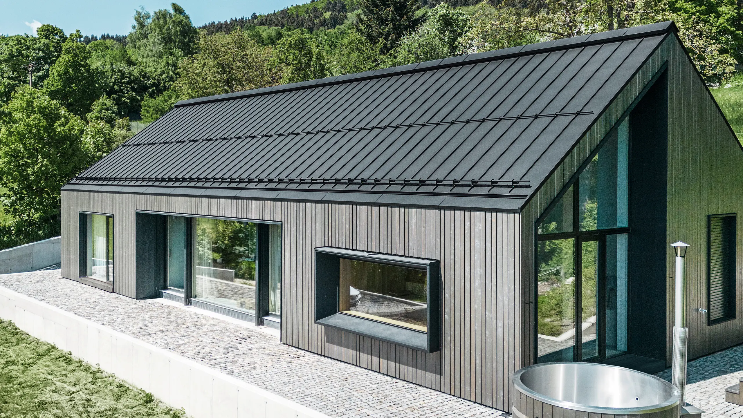 Ein modernes Holzmassivhaus in Kuncice (CZ) wurde mit großen Fensterflächen ausgestattet. Das dunkle Holzmassivhaus verfügt über ein Dachsystem aus PREFALZ in der Farbe P. 10 Schwarz. Im hinteren Außenbereich steht ein Whirlpool mit einer Holzverkleidung.