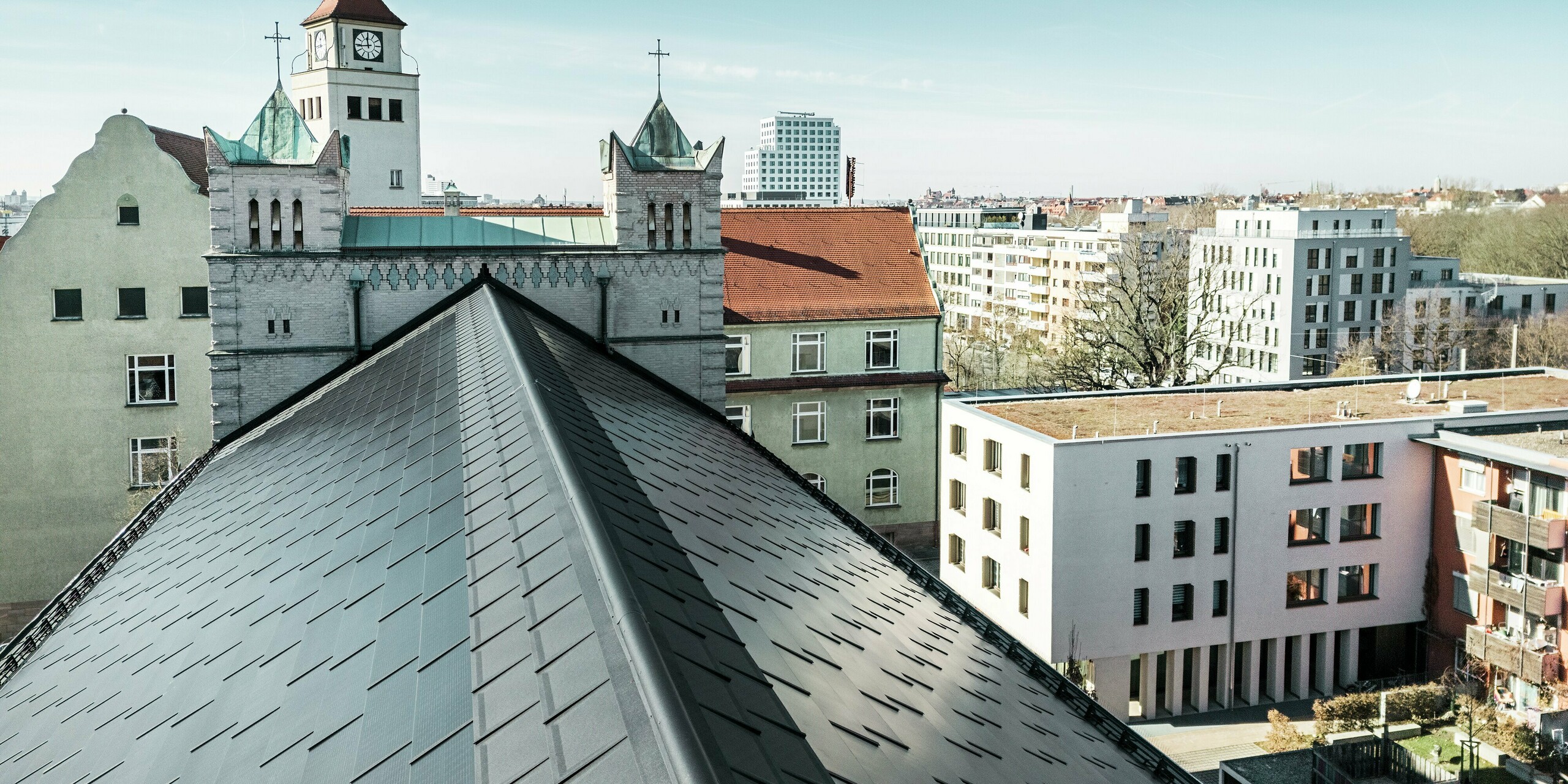 L'image montre la couverture précise de la toiture en aluminium de l'église Saint-Charles-Borromée de Nuremberg. Le toit de l'église a été équipé de tuiles solaires PREFA d'un noir élégant. La vue sur le faîtage marquant du toit à deux versants souligne l'intégration réussie de cette solution photovoltaïque moderne intégrée au bâtiment historique. Les tuiles solaires s'intègrent harmonieusement au bâtiment classé et alimentent l'église et plusieurs bâtiments voisins en énergie efficace et durable.