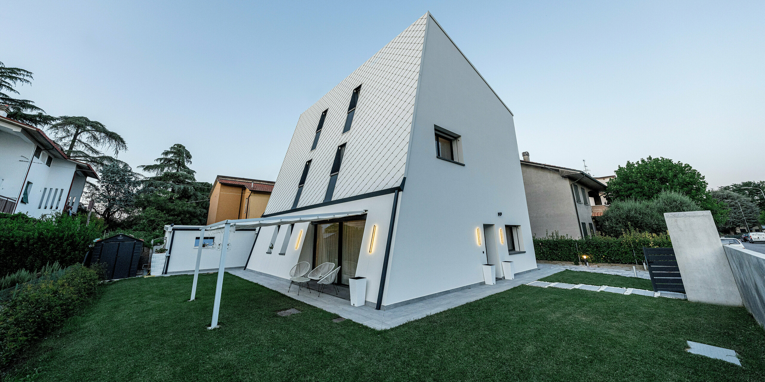 Abendliche Schrägansicht eines modernen Einfamilienhauses in Forlì, Italien, mit klarer, skulpturaler Architektur und großzügiger Grünfläche. Die geneigte Hauptfassade ist mit PREFA Wandraute 29 × 29 in P.10 Reinweiß gestaltet und wirkt durch das feine Aluminiumraster besonders plastisch im weichen Licht. Schlanke Fenster, die überdachte Terrasse und die ruhige Gartenszene unterstreichen den hohen Wohnkomfort. Die PREFA Aluminiumfassade überzeugt durch langlebige Qualität, Witterungsbeständigkeit und einen dauerhaft eleganten Auftritt.