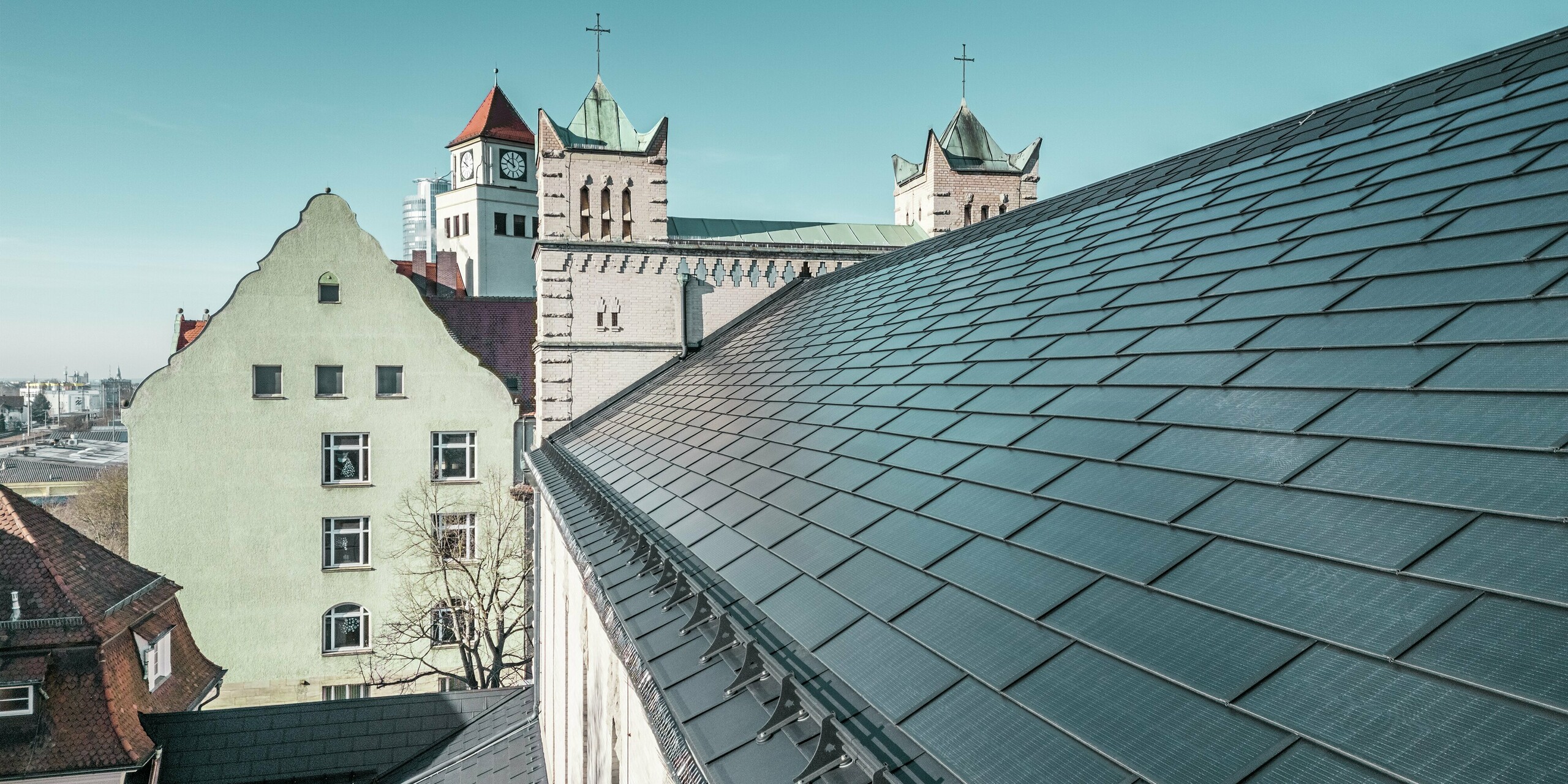Ce gros plan du toit de l'église Saint-Charles-Borromée de Nuremberg met en valeur les tuiles solaires PREFA, installées avec précision et d'un noir élégant. Ces modules solaires parfaitement intégrés s'intègrent harmonieusement à l'architecture historique et représentent une source d'énergie durable. En arrière-plan, on distingue les imposants clochers de l'église, avec leurs toits pointus classiques. La solution de toiture PREFA, en aluminium robuste, allie parfaitement préservation du patrimoine et énergie renouvelable.