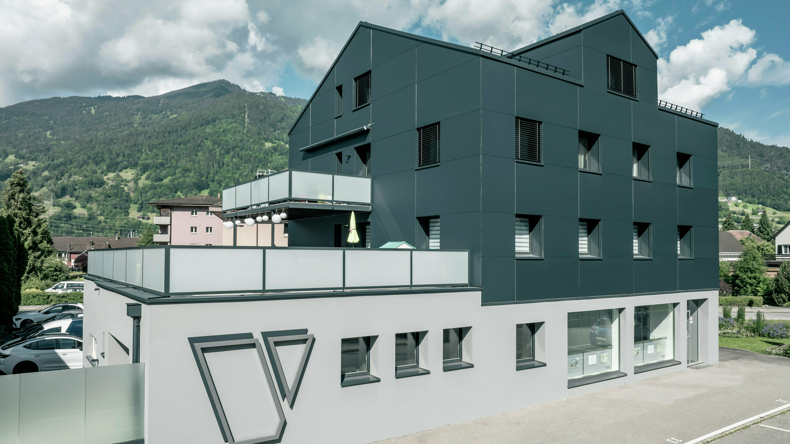 Le bureau d'architecture Voser à Landquart, dans le canton suisse des Grisons, avec une façade en aluminium PREFA en anthracite. Le bâtiment a été rénové et équipé de panneaux composites en aluminium PREFABOND. Le design allie des lignes claires et des matériaux de haute qualité. Les grands balcons avec parapets en verre et le logo frappant du cabinet d'architectes Voser sur le côté du bâtiment sont frappants. Un paysage de montagne verdoyant peut être vu en arrière-plan.