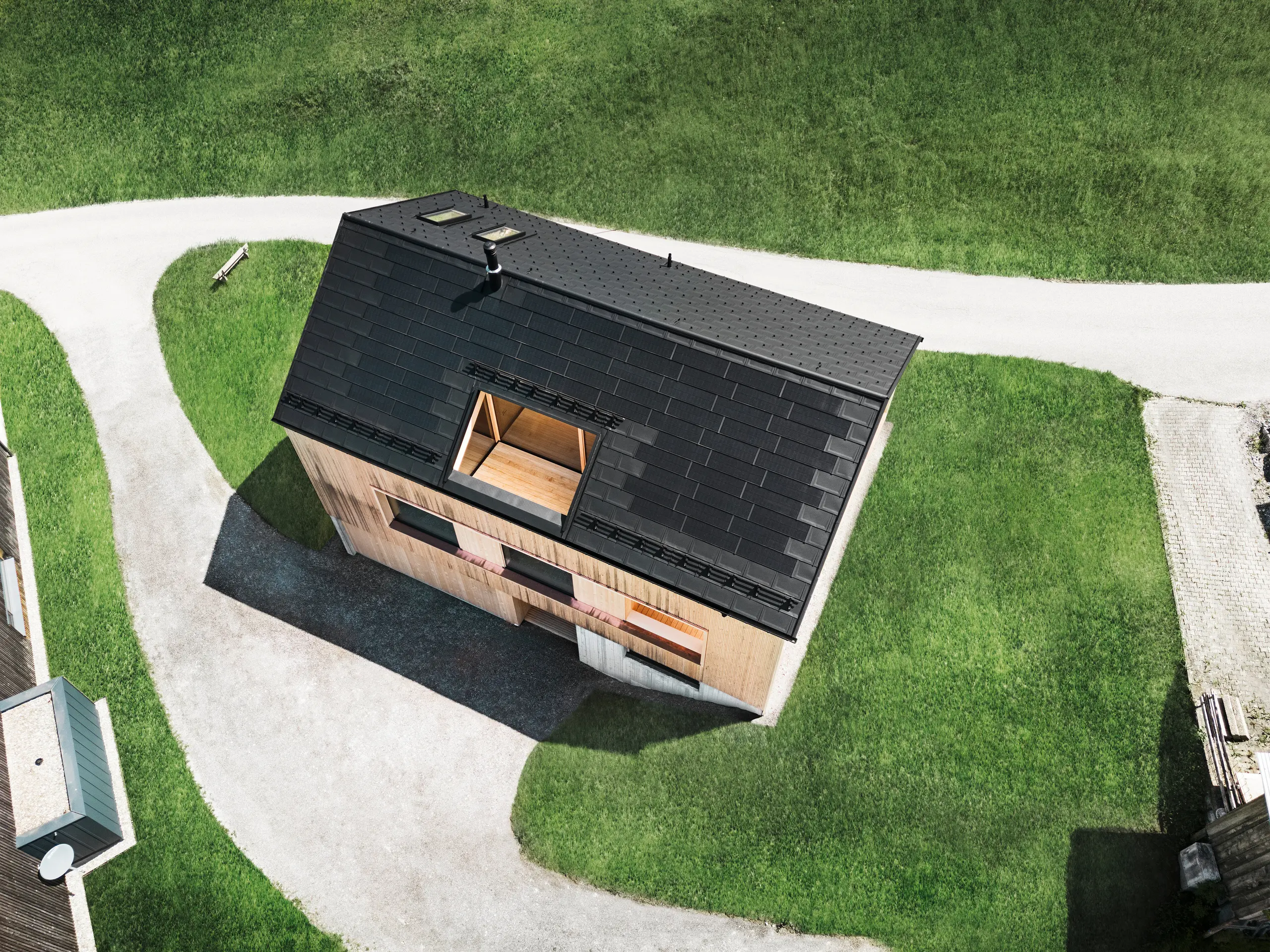 Drohnenaufnahme mit Blick von oben auf ein ökologisches Einfamilienhaus in Egg, das sich mit seinem schwarzen PREFA Solardach und den traditionellen PREFA Dachplatten R.16 von den umliegenden grünen Rasenflächen abhebt. Die strategisch angeordneten Solarmodule maximieren die Energieeffizienz, während die klare Geometrie des Daches und die natürlich wirkende Holzfassade eine Verbindung zwischen modernem Wohnen und der ländlichen Umgebung schaffen. Dieses Haus verkörpert eine fortschrittliche, umweltbewusste Bauweise, die sowohl funktional als auch optisch beeindruckt.