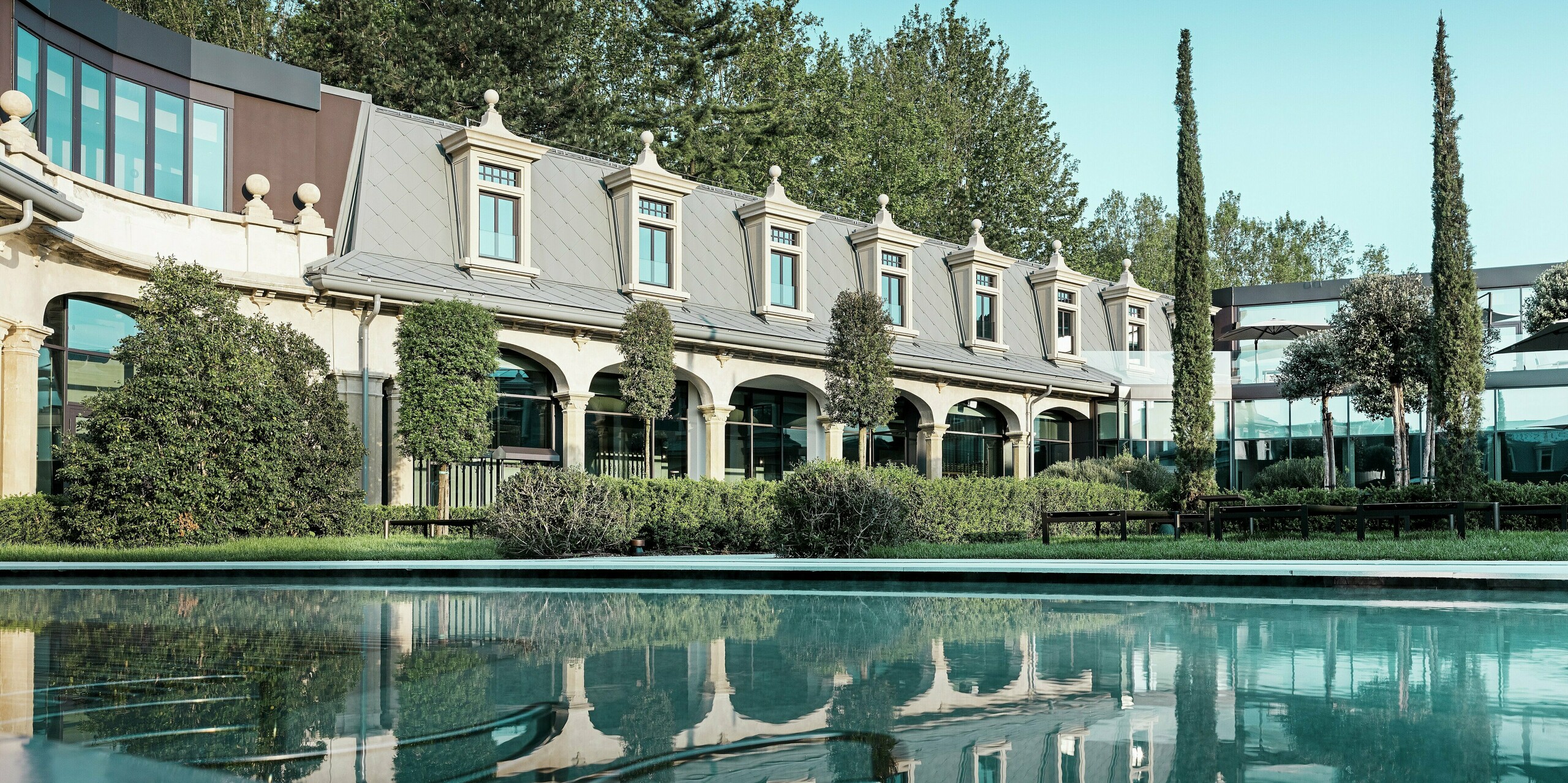Vue sur le bassin paisible de la façade historique des thermes « De Montel – Terme Milano ». La toiture remarquable, composée de losanges PREFA 44 x 44 P.10 gris souris et de lucarnes finement travaillées, se reflète de manière impressionnante dans l'eau. Entouré d'espaces verts soignés, de cyprès et de structures en verre modernes, le bâtiment offre une harmonie parfaite entre tradition, nature et architecture contemporaine.