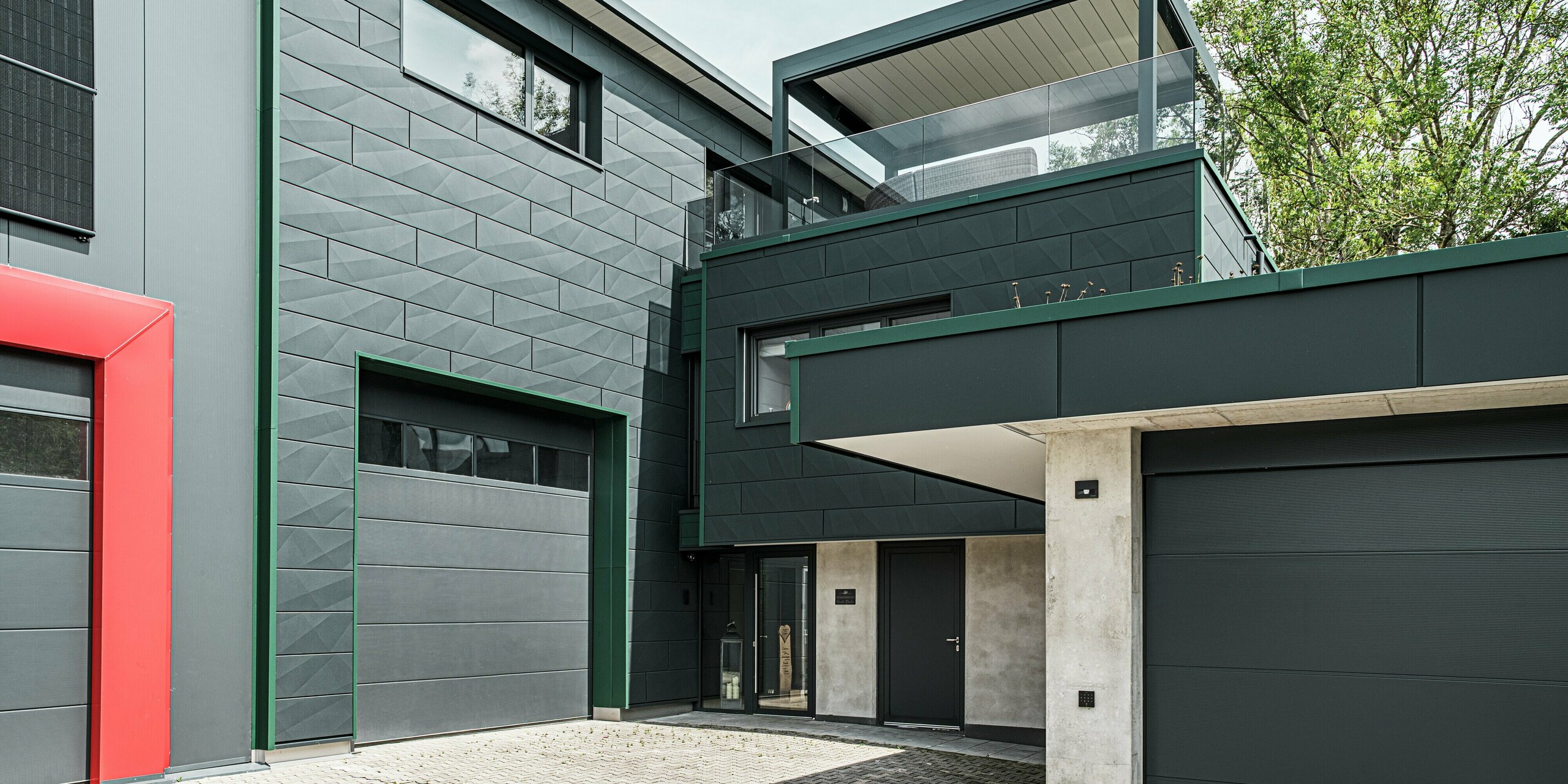 Das moderne Einfamilienhaus in Türkheim besticht durch seine markante Architektur und seine auffällige Fassadengestaltung. Auf dem Bild sind PREFA Siding X-Paneele in der Farbe P.10 Anthrazit zu sehen. Die Leisten an den Gebäudeecken sind in der Farbe P.10 Moosgrün gehalten. Die Siding-X-Fassade verleiht dem Gebäude eine elegante und dynamische Optik. Besonders auffällig ist der rote Torrahmen im Vordergrund, der die Garage markiert und einen starken Kontrast zur Fassade bildet. Die Dachterrasse über dem Eingangsbereich mit ihrer modernen Glasbrüstung sorgt für einen offenen, luftigen Eindruck und gibt den Blick auf den klaren Himmel frei. Die Aluminiumfassade überzeugt zudem mit Langlebigkeit und Wetterbeständigkeit.