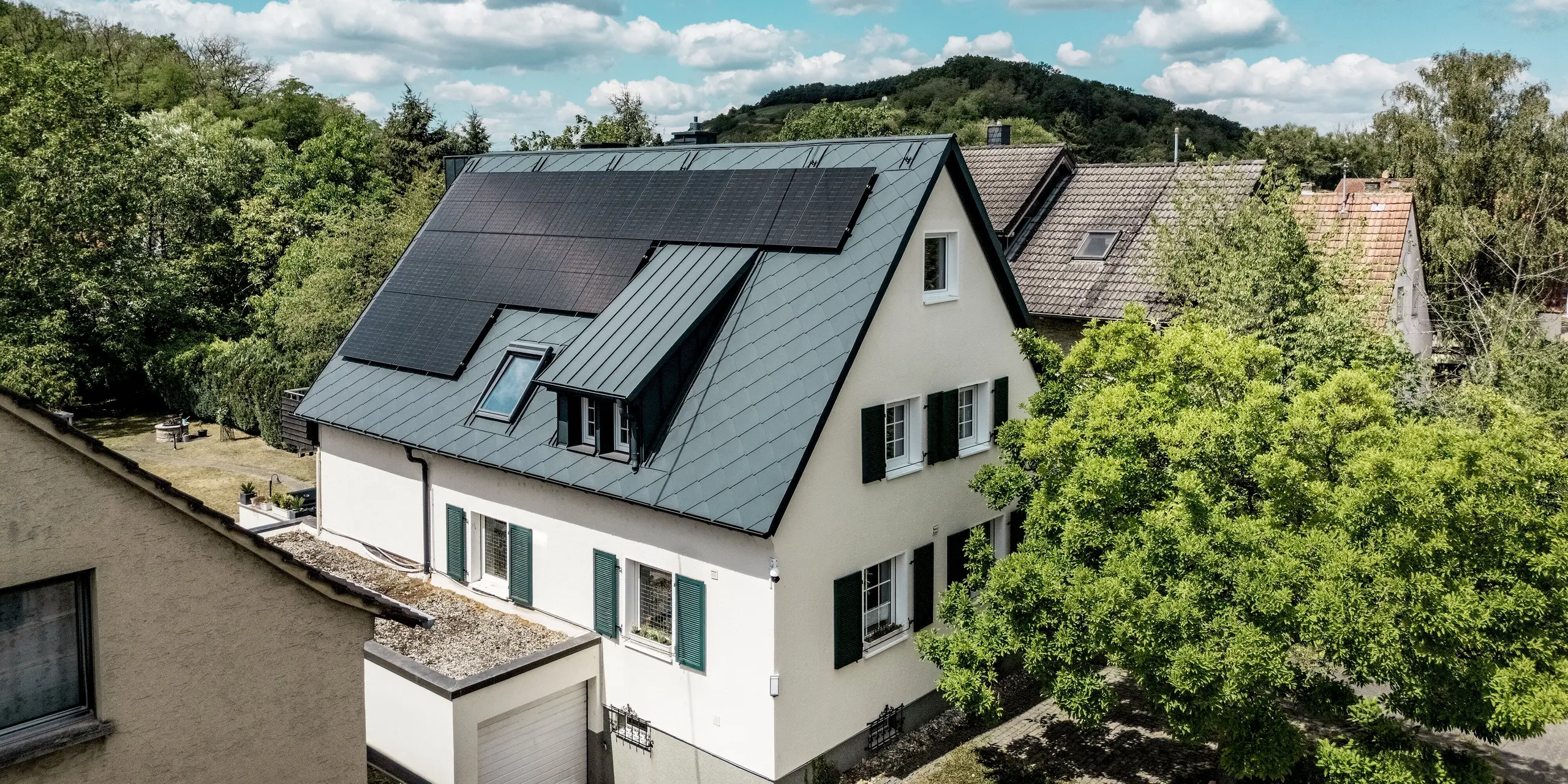 Luftaufnahme eines sanierten Einfamilienhauses in Hargesheim mit markantem PREFA Aluminiumdach aus Dachrauten 44 × 44 in P.10 Anthrazit. Großflächige Photovoltaikmodule, befestigt mit dem PREFA Solarmontagesystem, und eine mit PREFALZ verkleidete Dachgaube prägen das moderne Erscheinungsbild. Die leichte, langlebige und witterungsbeständige Dacheindeckung bildet einen eleganten Kontrast zur hellen Putzfassade.
