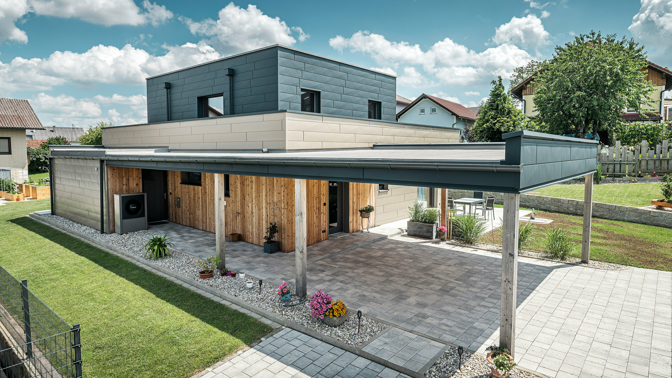 Luftaufnahme der Zufahrt eines modernen Einfamilienhauses in Wippenham, Österreich, mit einer zweifarbigen PREFA Aluminiumfassade in Bronze und Anthrazit. Das Haus zeigt die stilvolle Integration von Siding.X Fassadenpaneelen und Holzverkleidung. Deutlich sichtbar ist der überdachte Carport, der nahtlos in die Architektur des Hauses integriert ist. Der gepflegte Garten und die Terrassenfläche unterstreichen das moderne und funktionale Design des Gebäudes.