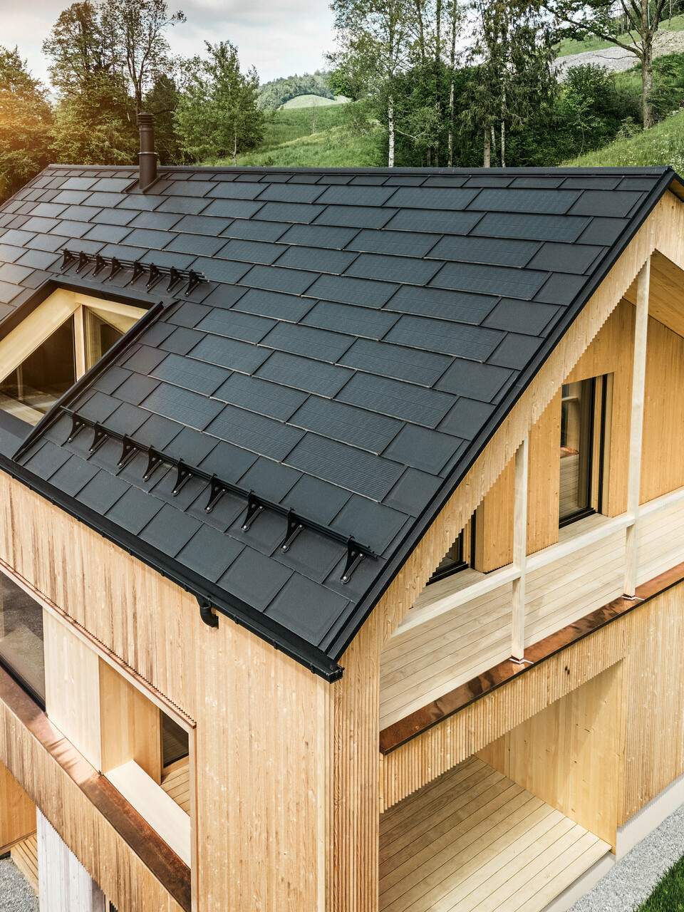 Blick auf ein modernes, nachhaltiges Holzhaus in Egg mit einem PREFA Solardach, das durch seine dunkle, strukturierte sowie elegante Erscheinung auffällt. Die Solarmodule sind strategisch zwischen den klassischen Dachplatten R.16 platziert, um eine optimale Sonneneinstrahlung zu nutzen und sich dabei ästhetisch in das Gesamtdesign einzufügen. Das Haus steht im Einklang mit seiner grünen Umgebung, während die warme Abendsonne die natürliche Holztextur der Fassade unterstreicht und die solide Bauweise betont. Ein harmonisches Beispiel für umweltbewusstes Bauen und Energieeffizienz.