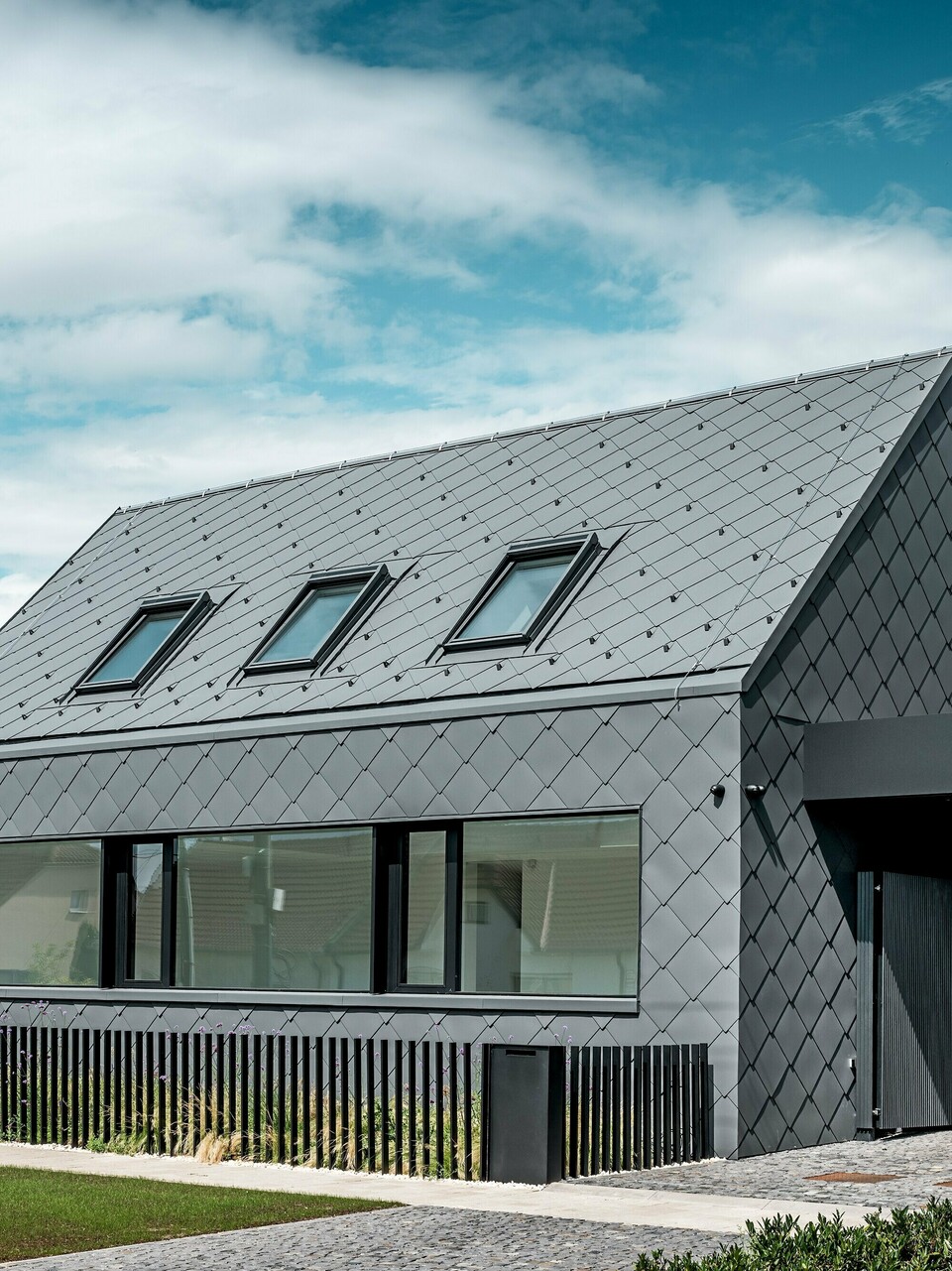 Modernes Einfamilienhaus in Trnava‑Modranka mit Aluminiumhülle: Die gesamte Gebäudehülle dieses schlichten Gebäudes wurde mit PREFA Dach- und Wandrauten 44 × 44 in P.10 Dunkelgrau gestaltet. Die markante Rautenstruktur zieht sich nahtlos über Dach und Fassade und verleiht dem Haus eine starke, monolithische Wirkung. Große Fensteröffnungen setzen klare architektonische Akzente, während das witterungsbeständige PREFA Aluminium langlebigen Schutz und eine edle, matte Optik bietet. Der helle Himmel verstärkt den Kontrast zur dunklen Gebäudehülle und betont das moderne Design.