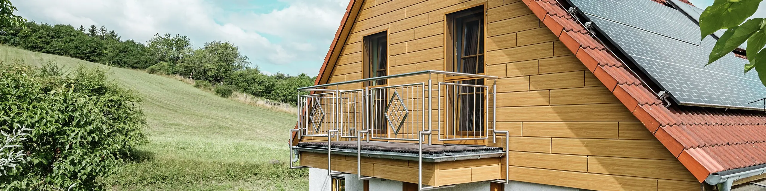 La photo montre une charmante maison individuelle à Blieskastel, dont le pignon fraîchement rénové avec des Sidings PREFA couleur chêne naturel, attire immédiatement le regard. L'aspect bois de l'aluminium, plus durable, s'harmonise parfaitement avec le toit en tuiles rouges et la vaste véranda. La verdure luxuriante et le parterre de fleurs soigneusement aménagé forment une scène idyllique et vivante. La rénovation de façades avec PREFA confère à la maison un aspect naturel et en impose par sa robustesse : résistance aux intempéries, fidélité aux couleurs d'origine et longue durée de vie, pratiquement sans entretien.
