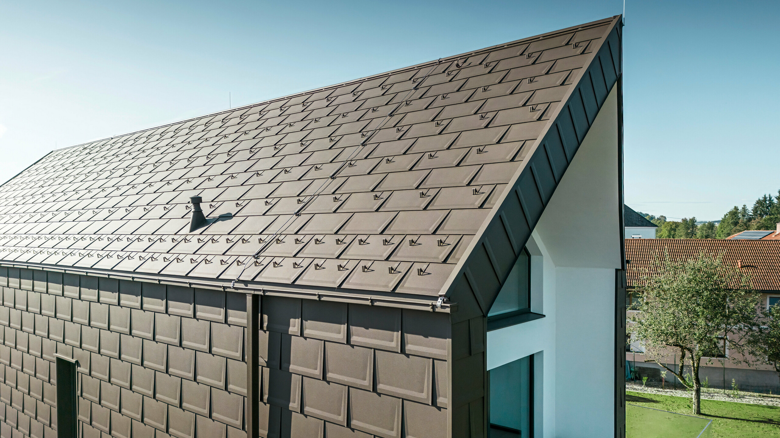 Een perspectivisch aanzicht van het dak en de gevel van een moderne eengezinswoning in Neukirchen, Oostenrijk. De PREFA R.16 dakpan in bruintint P.10 biedt een esthetische en functionele dakbedekking die perfect past bij het architectonische ontwerp van de woning. De scherpe nok van het dak en de nauwkeurige plaatsing van de dakpanelen tonen de kwaliteit van PREFA. De kleine formaten aluminium producten passen perfect bij de wit gepleisterde gevel en het omringende groene landschap.