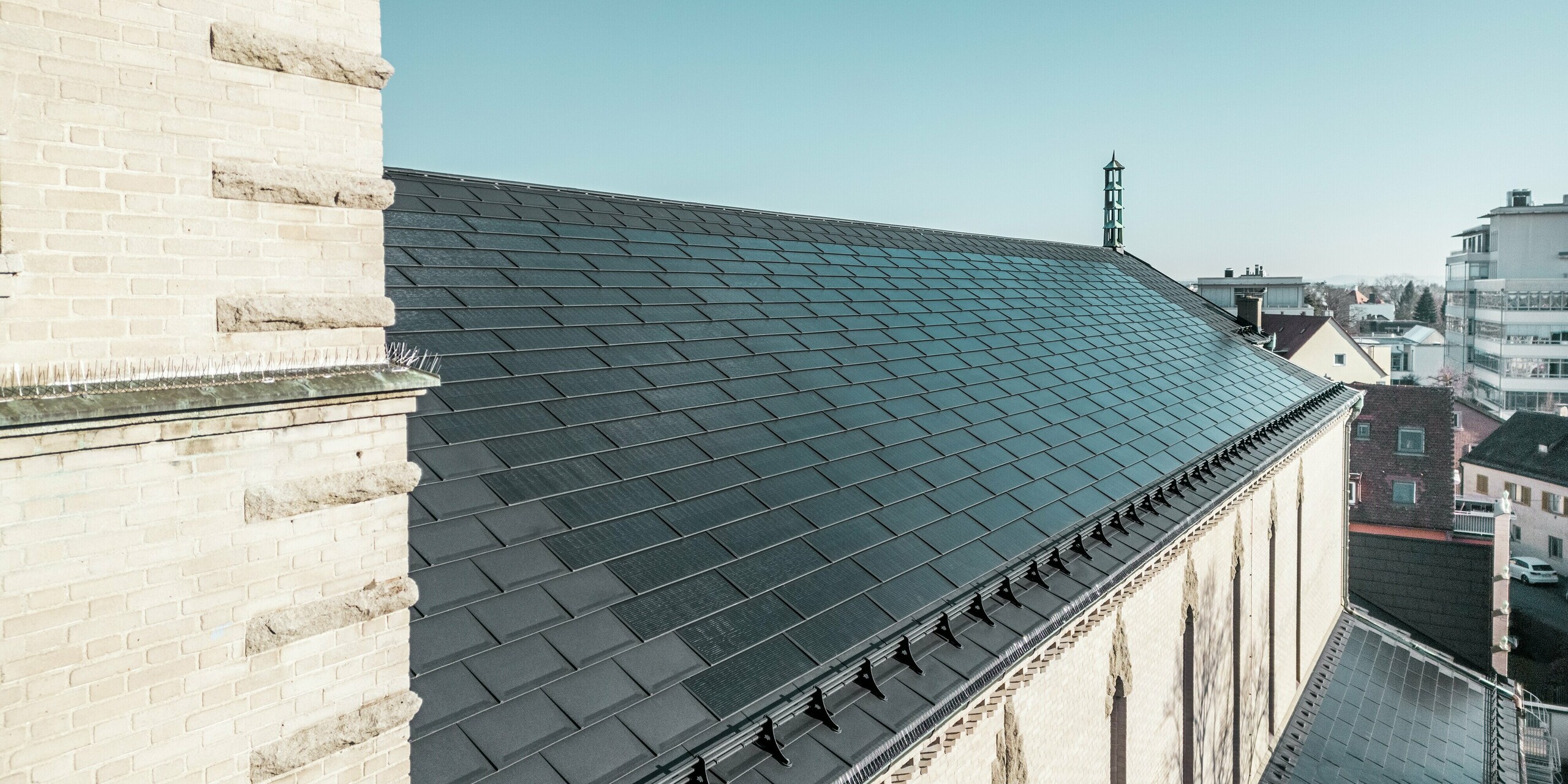 La vue au-delà du clocher révèle les tuiles solaires PREFA d'un noir élégant sur le toit de l'église Saint-Charles-Borromée de Nuremberg. La perspective met en valeur l'intégration parfaite des modules solaires, qui s'intègrent harmonieusement au toit à pignon du bâtiment classé. Les éléments décoratifs marquants du mur de l'église, qui soulignent l'architecture expressionniste franconienne, sont également visibles. Les tuiles solaires fournissent une énergie durable à l'église et aux bâtiments environnants et constituent simultanément une solution esthétique dans le domaine de la préservation du patrimoine historique.