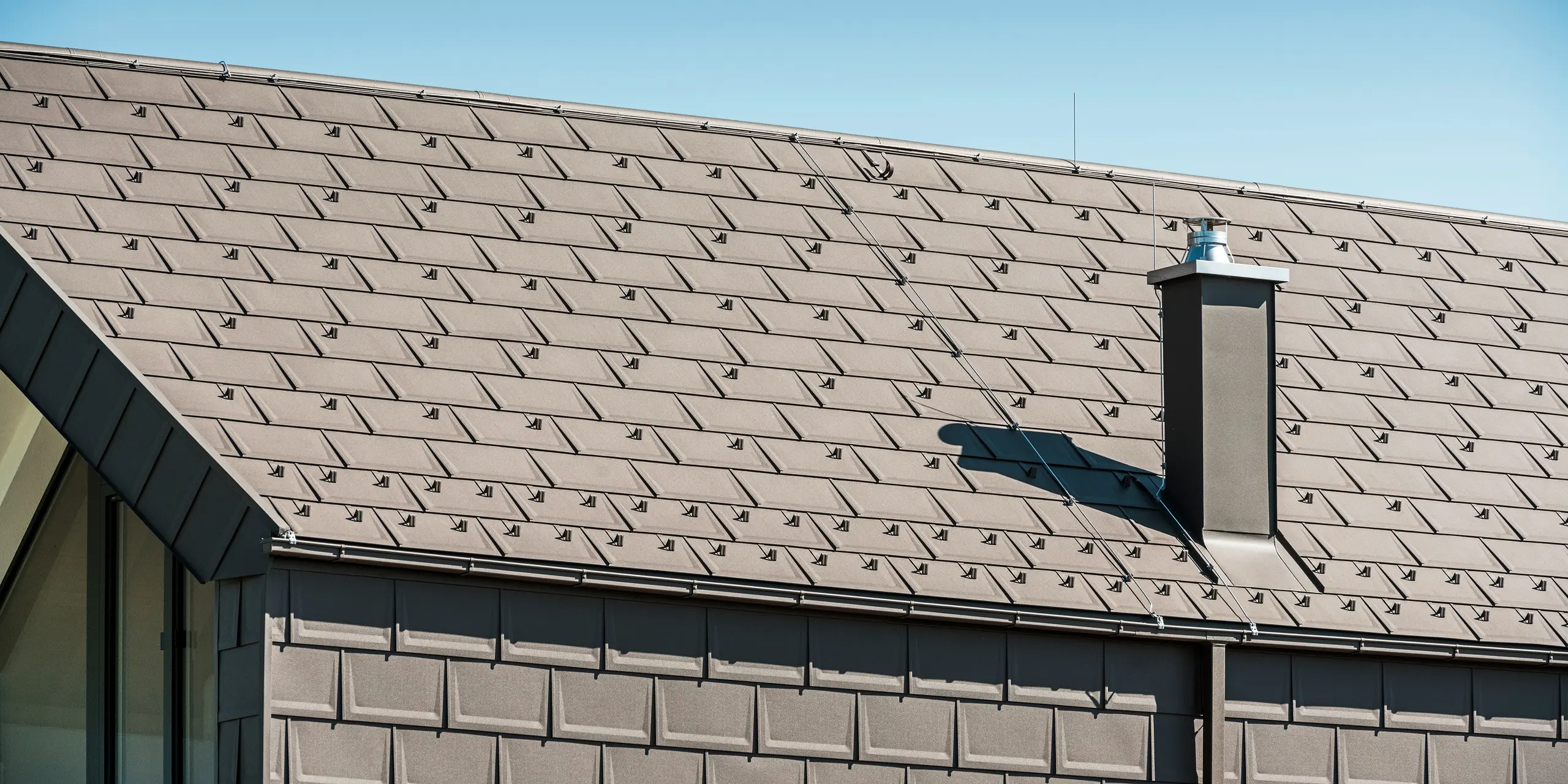 Seitenansicht eines Hauses in Neukirchen, Österreich, mit PREFA R.16 Dachplatten in P.10 braun auf Dach und Fassade. Der Dachaufbau zeigt die handwerkliche Qualität und Langlebigkeit der PREFA Produkte. Ein markanter Kamin und eine moderne Dachentwässerung in Form einer Kastenrinne und Vierkantrohren ergänzen das Dachsystem und unterstreichen die klare Linienführung und durchdachte Gestaltung des Gebäudes.