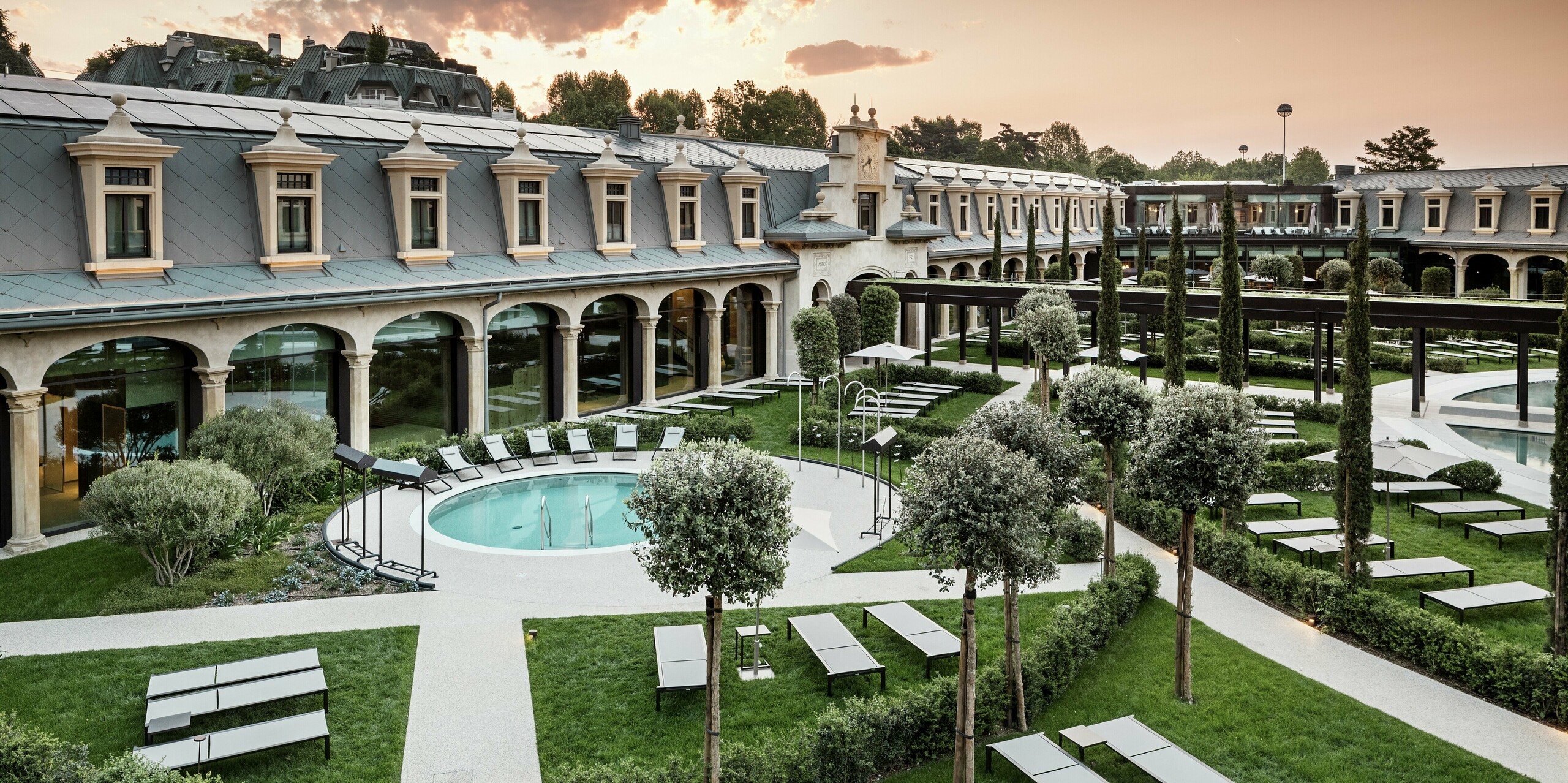 Ambiance nocturne au parc thermal « De Montel – Terme Milano » avec une vue imprenable sur la façade Art nouveau classée. La toiture en losanges PREFA 44 × 44 P.10 gris souris allie durabilité et esthétique. Au premier plan, pelouses, sentiers bien entretenus, oliviers et piscine thermale circulaire invitent à la détente.