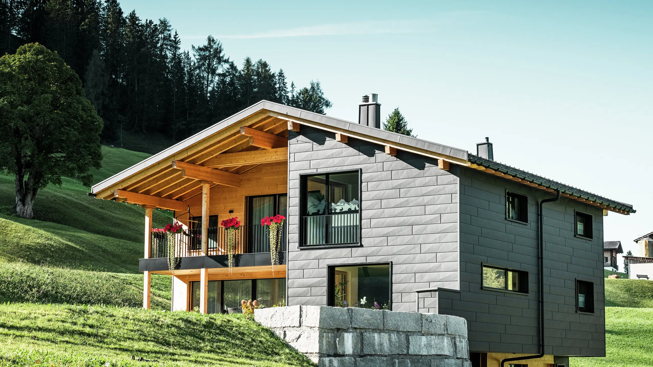 Cette maison individuelle moderne à Ascharina, en Suisse, s'intègre harmonieusement dans le paysage alpin verdoyant. Le bâtiment est entièrement revêtu de panneaux de façade PREFA Siding.X durables et résistants aux intempéries en P.10 noir, qui confèrent à la structure une apparence unique. Le balcon en bois complète parfaitement la coque en aluminium foncé et apporte une touche naturelle.